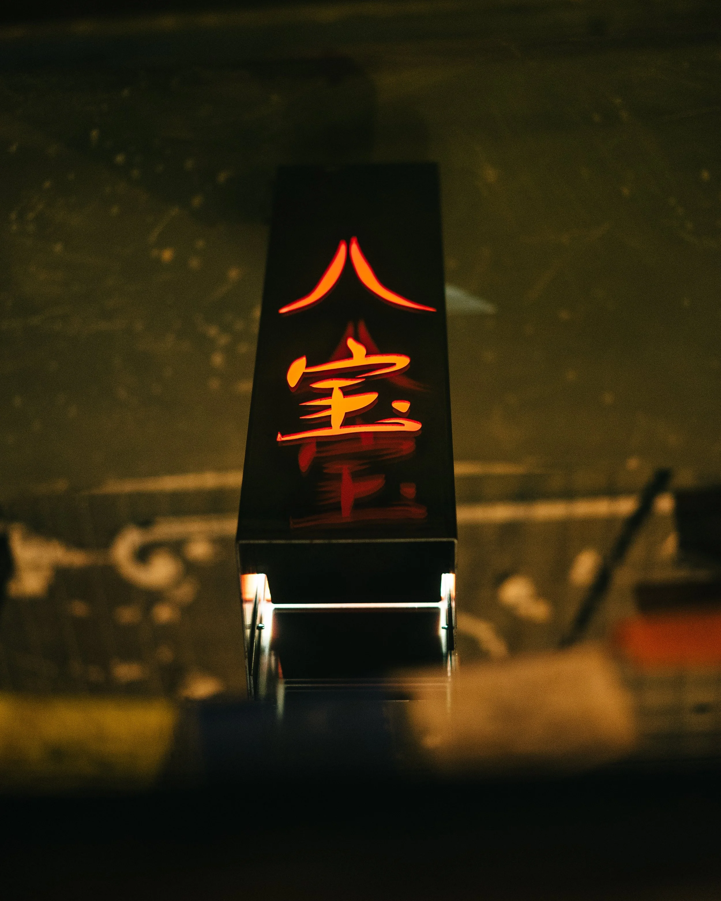Illuminated sign with red Chinese characters on a black background, reflected underneath, hanging against a dark wall.