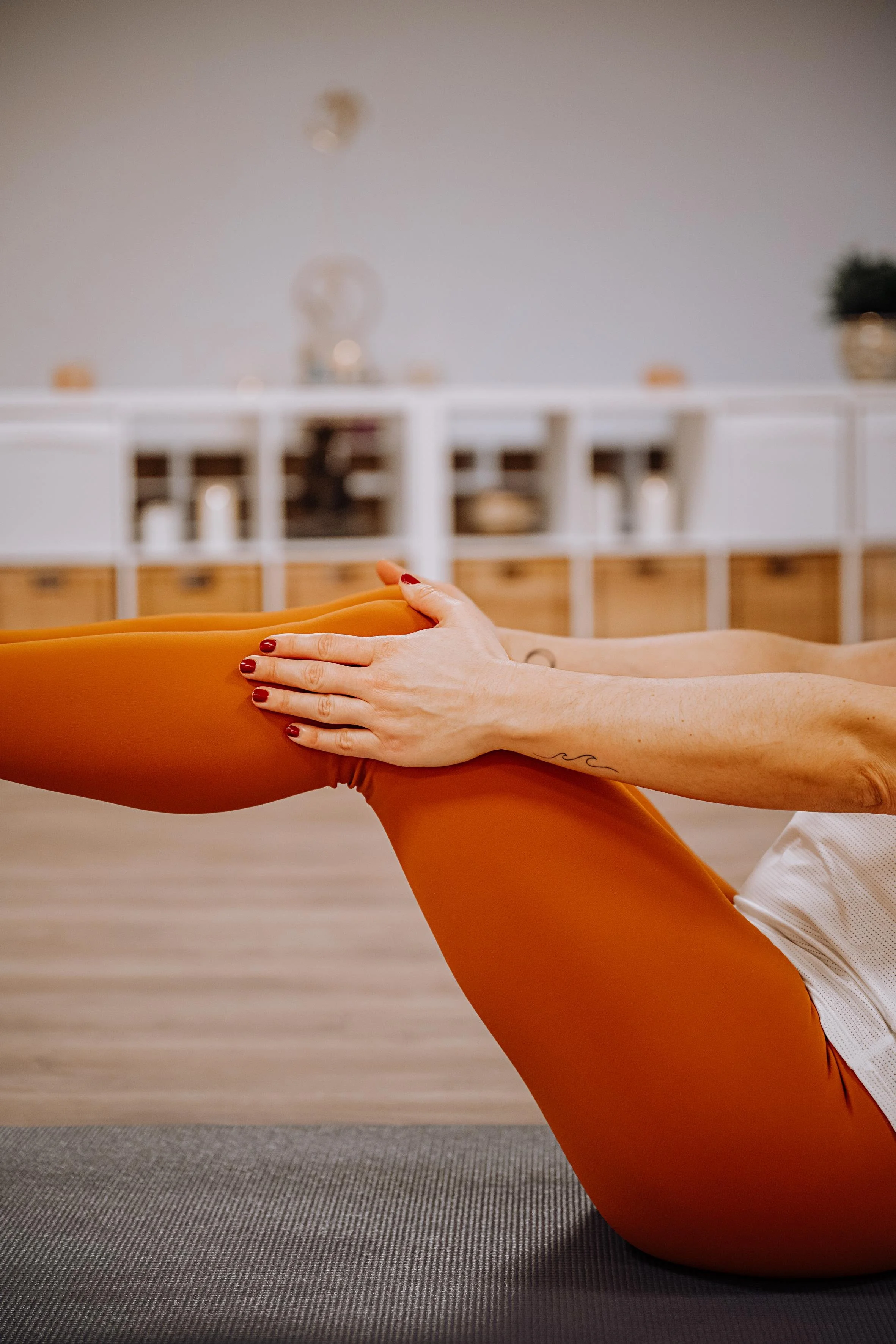 Person, die Yoga macht, hält ihr Bein mit beiden Händen im Innenbereich, auf einer Yogamatte in einem Raum mit weißem und Holzschrank im Hintergrund.