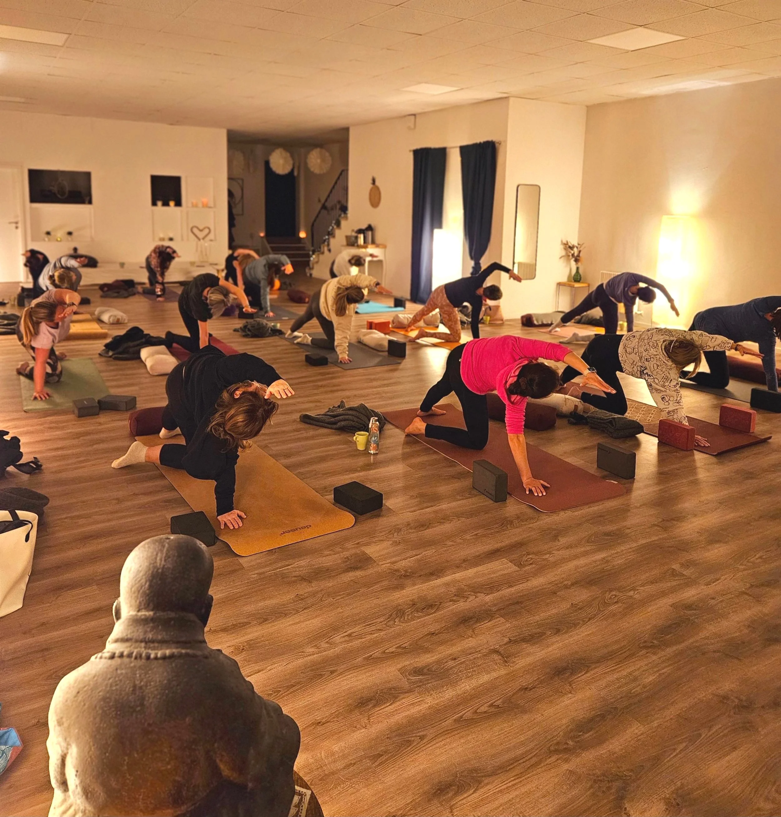 Menschen machen Yoga in einem warm erleuchteten Raum auf Yogamatten, mit Blöcken und Decken, während ein Lehrer im Vordergrund sitzt und zuschaut.