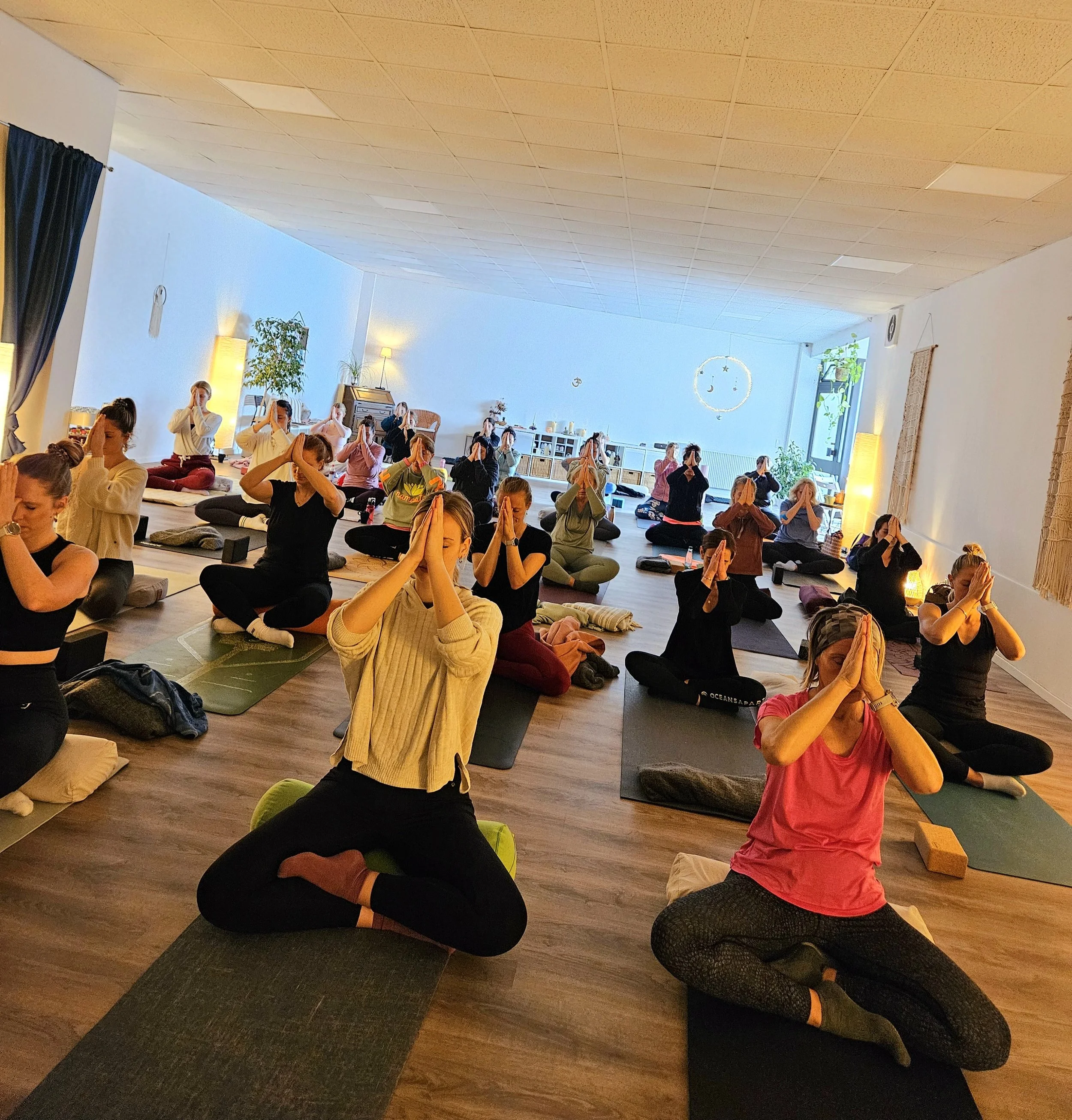 Viele Menschen sitzen in einem Yoga-Kurs, in gekreuzten Beugen, mit Händen vor der Brust in Gebetshaltung, in einem hell erleuchteten Raum.