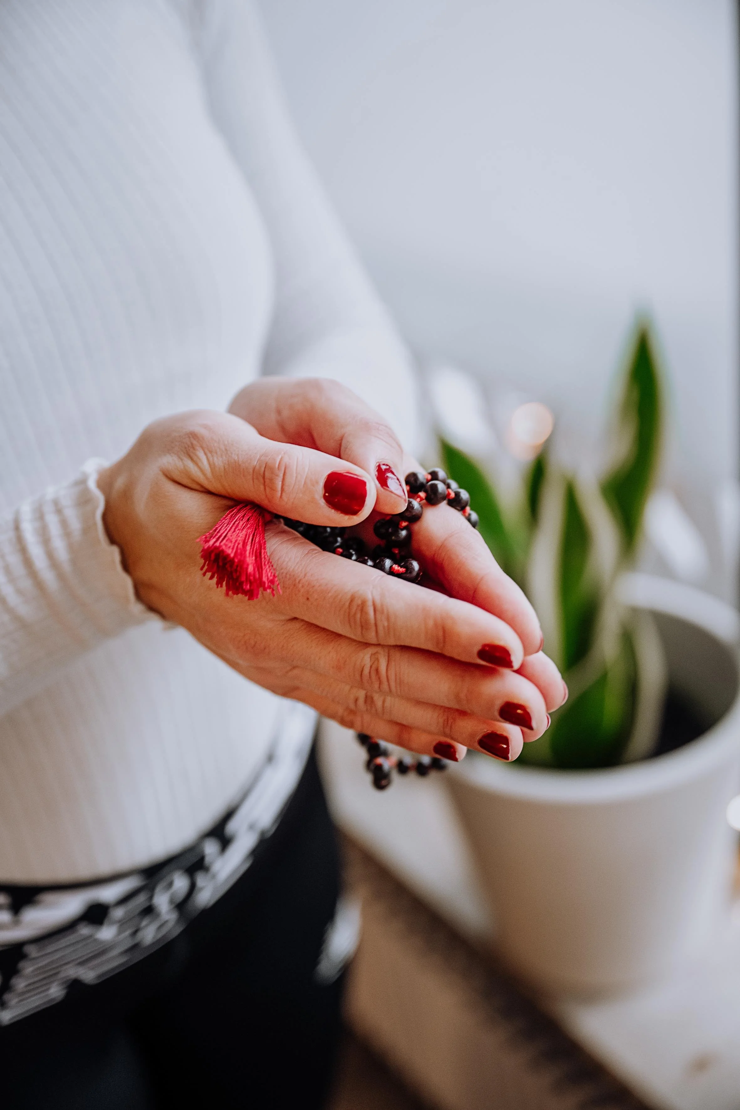 Eine Frau mit roten Nagellack hält eine Perlenkette mit einer roten Quaste in den Händen, im Hintergrund eine Topfpflanze.