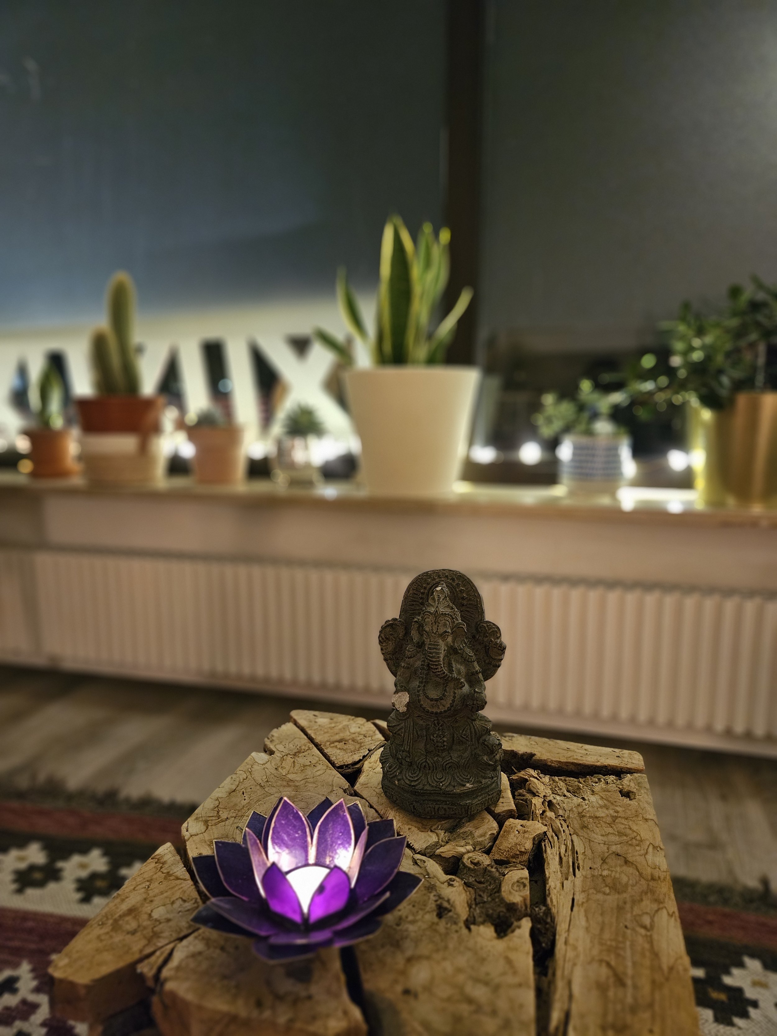 Ein kleiner Hindu-Götterfigur auf einem Holztisch, mit einer leuchtenden Lotus-Flammenlampe davor, im Wohnzimmer mit Pflanzen im Hintergrund.