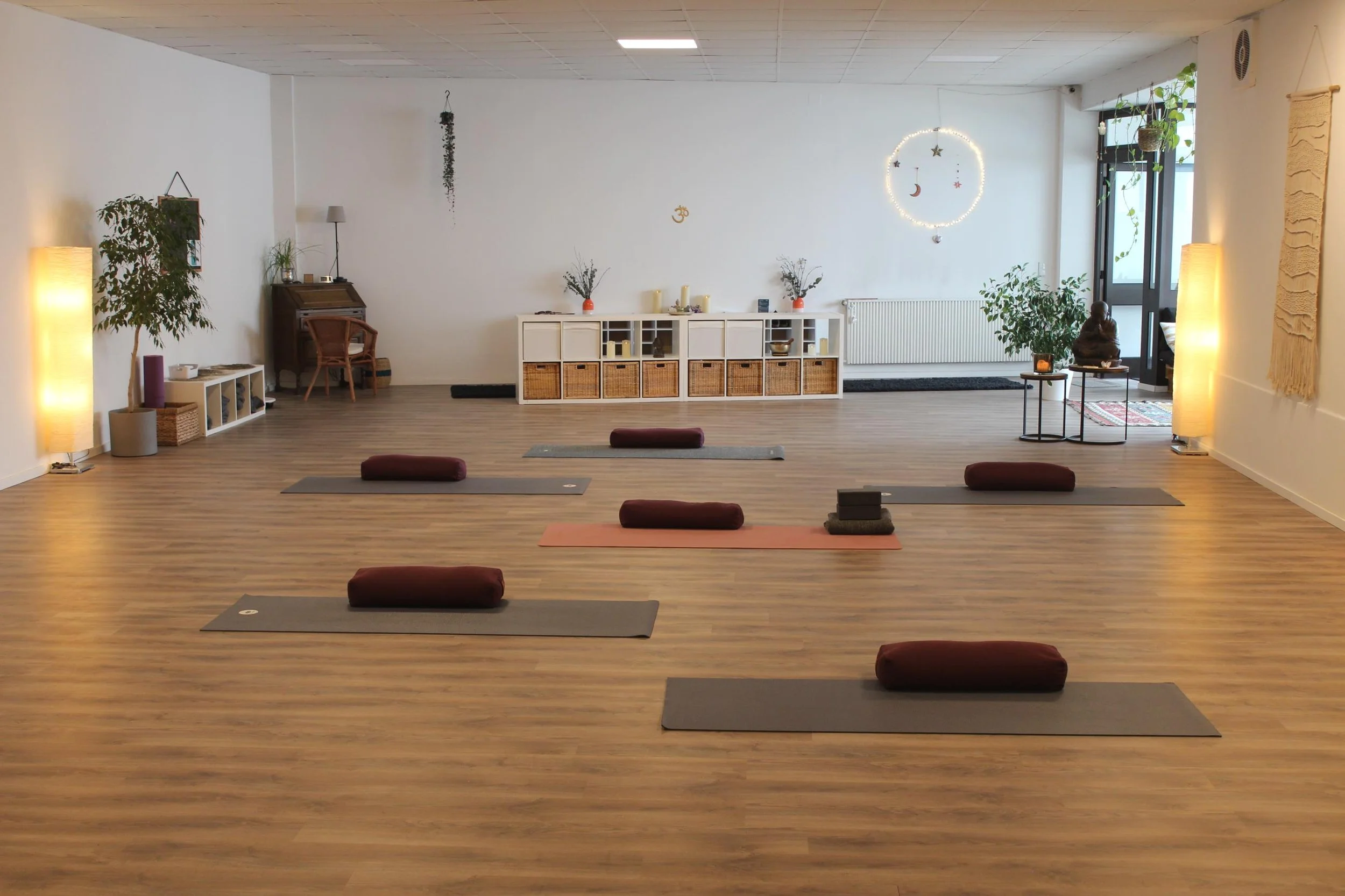 Ein Yogaraum mit sieben liegenden Matten, roten Bolstern und meditierenden Kissen, dekoriert mit Pflanzen, Laternen und einer Wanddekoration, bereit für eine Yogastunde.