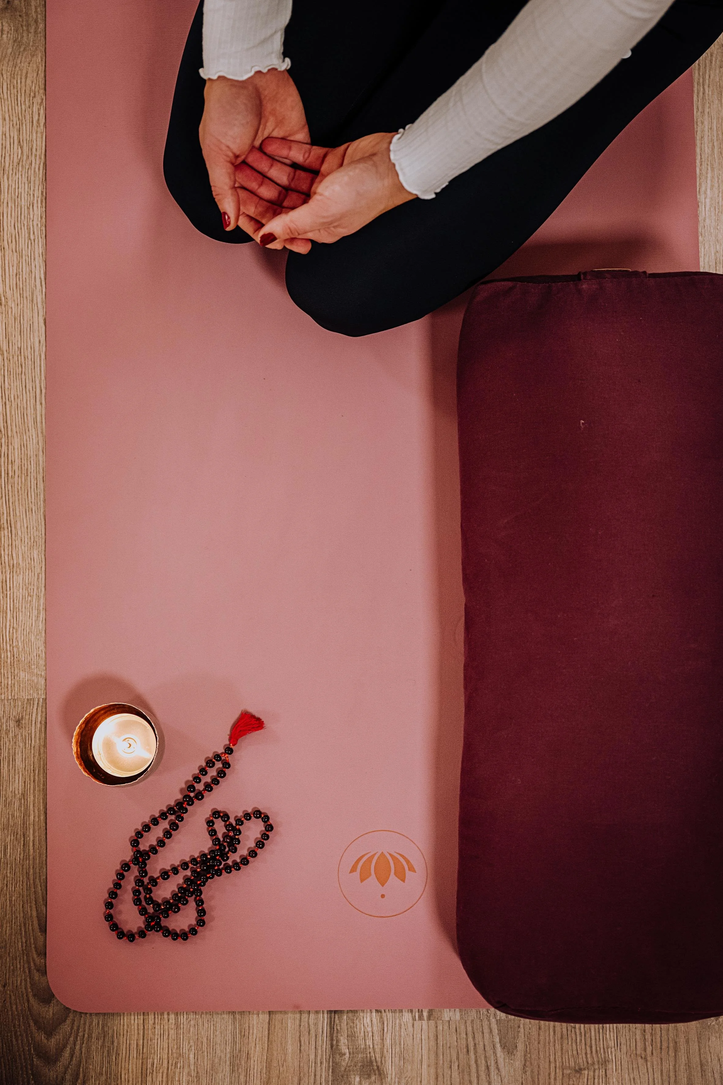 Eine Person sitzt im Schneidersitz auf einer Yoga-Matte, die auf einem Holzboden liegt, mit einer Kerze, einer Gebetskette und einem Logo auf der Matte.