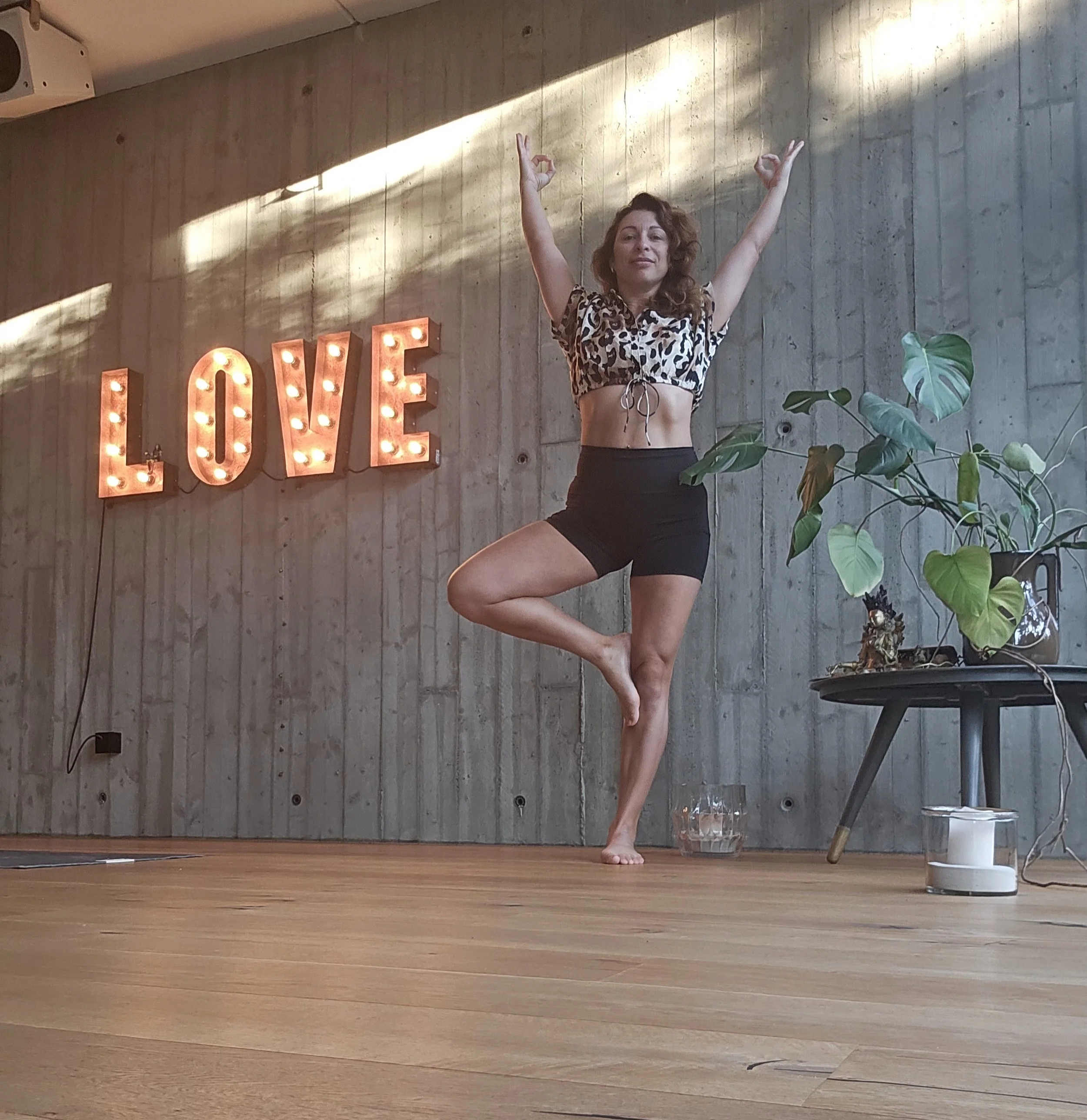 Frau führt Yoga-Übung im Raum mit Holzboden und einer Wand mit einer Leuchtdekoration, die das Wort Liebe zeigt, aus, während sie auf einem Bein balanciert und die Arme in der Luft ausstreckt.