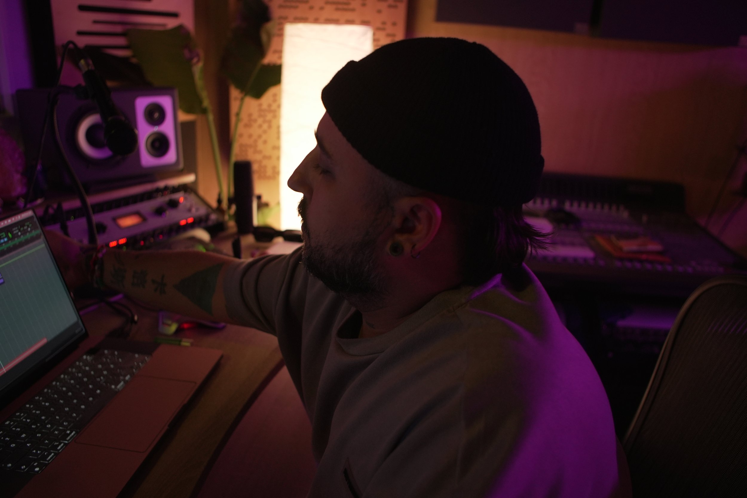 Un hombre en un estudio de grabación trabajando con una laptop y equipo de audio, iluminación tenue con luces moradas.