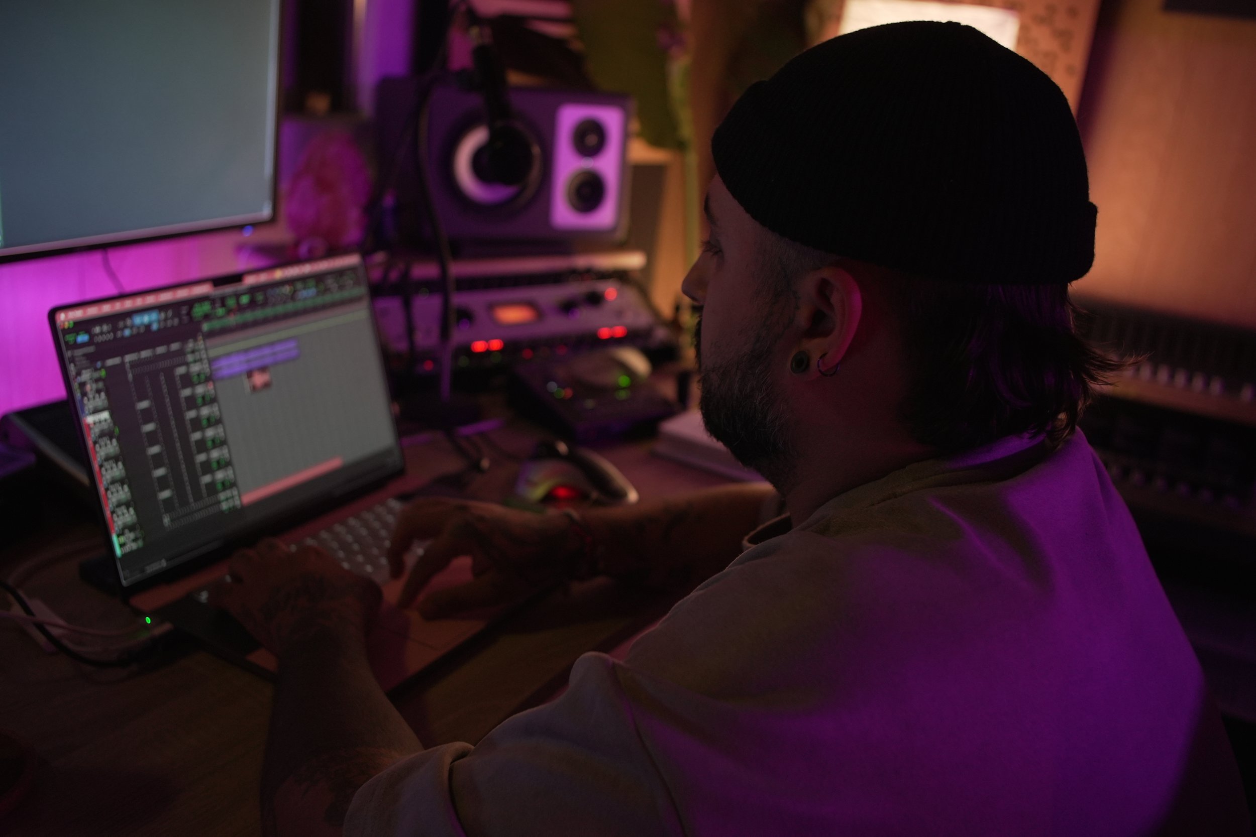 Persona trabajando en un estudio de grabación con equipo musical y computadora en un ambiente con luz morada y naranja.