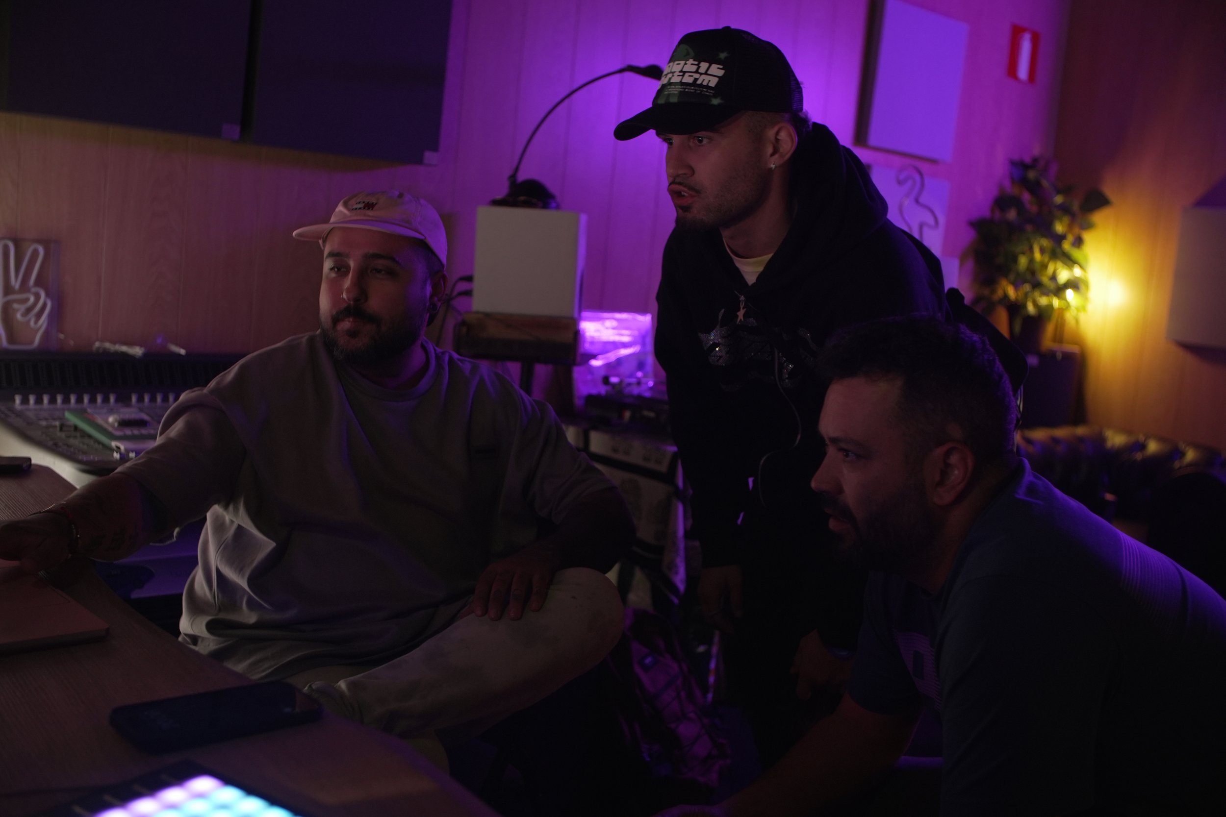 Tres hombres en un estudio de grabación con iluminación tenue y luz morada, uno sentado y dos de pie, supervisando o trabajando en un proyecto.