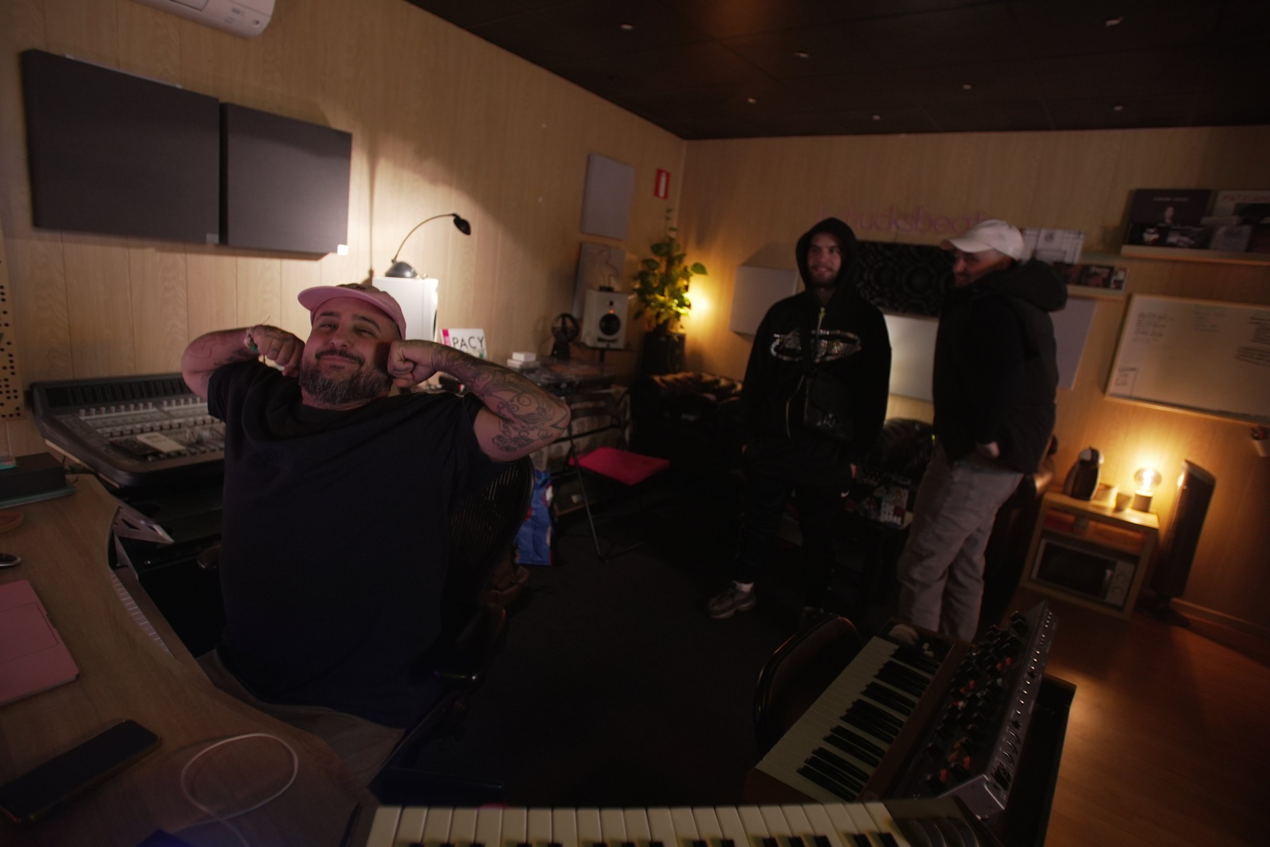Tres hombres en estudio de grabación, uno con gorra rosada y tatuajes en brazos, sonriente y con brazos flexionados, y dos más de pie, uno con capucha y el otro con gorra blanca.