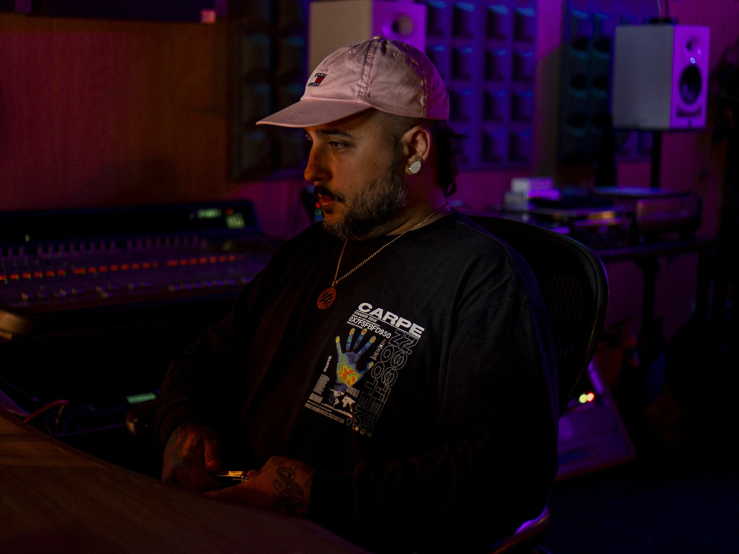 Joven en estudio de grabación, sentado frente a equipo de audio, con gorra rosa y ropa negra, ambiente con luces moradas y rojas.