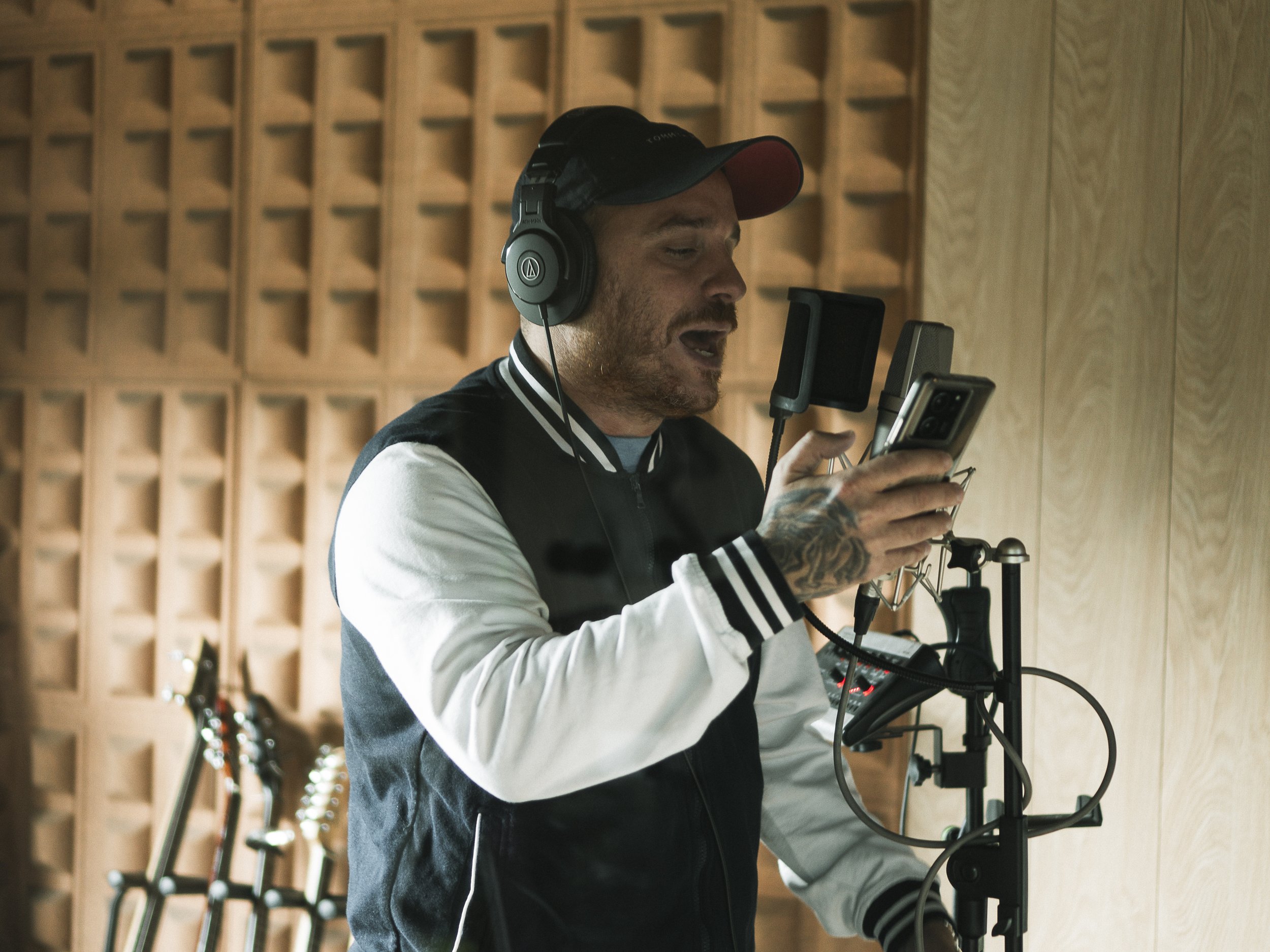 Hombre en estudio de grabación usando auriculares, sosteniendo un teléfono y cantando frente a un micrófono con una cabina.