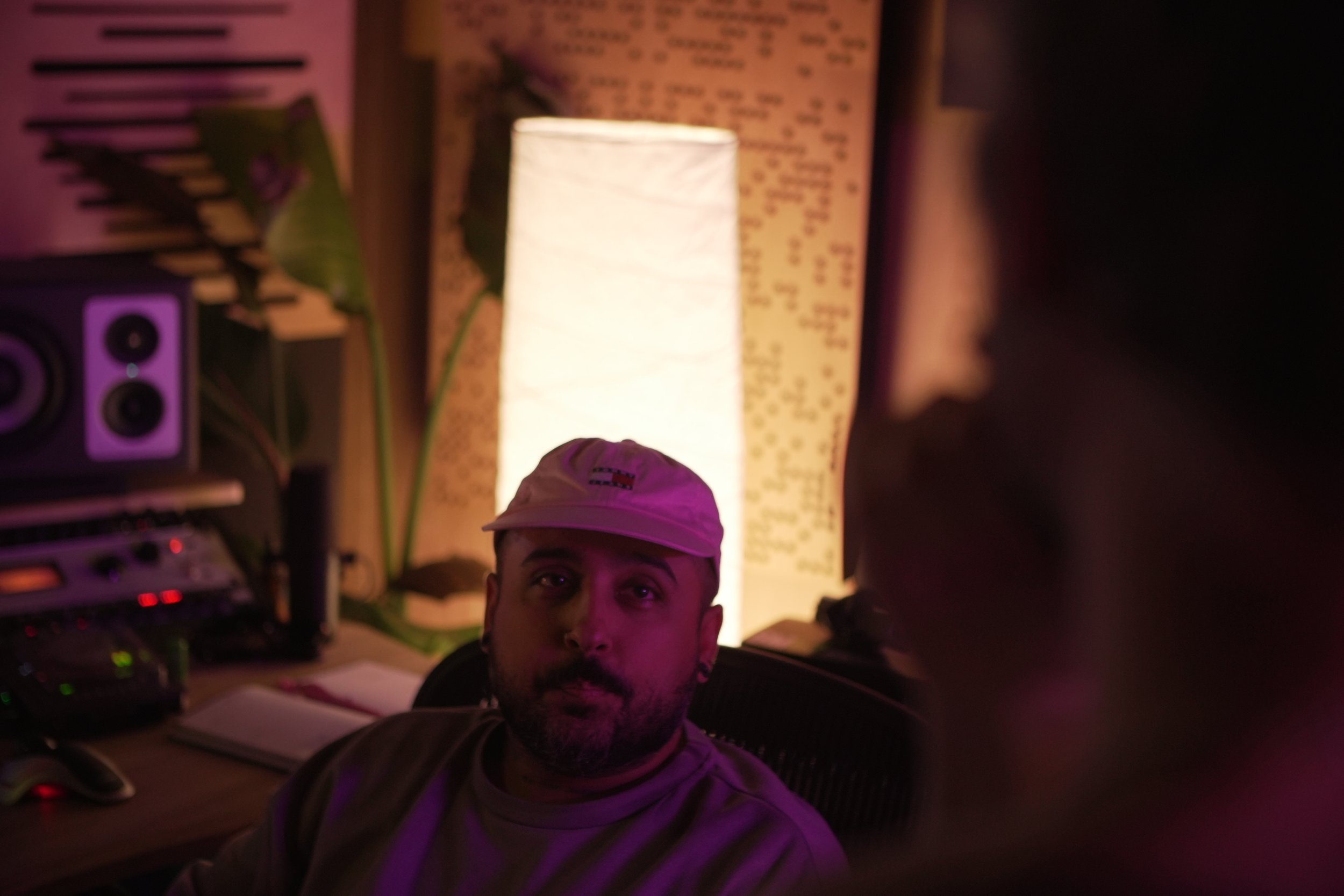 Retrato de un hombre con gorra en un estudio con luces ambientales y equipo de audio, con una lámpara de papel en el fondo.