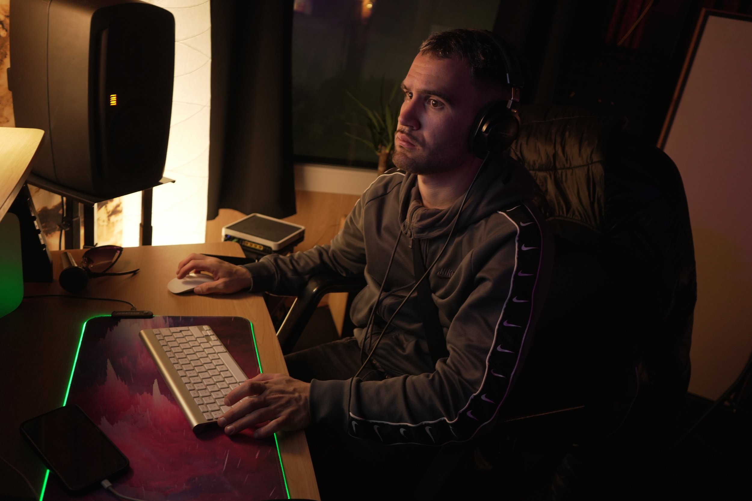 Hombre en una sala de estudio en la noche, usando audífonos y trabajando en una computadora con teclado retroiluminado en un ambiente de iluminación tenue.