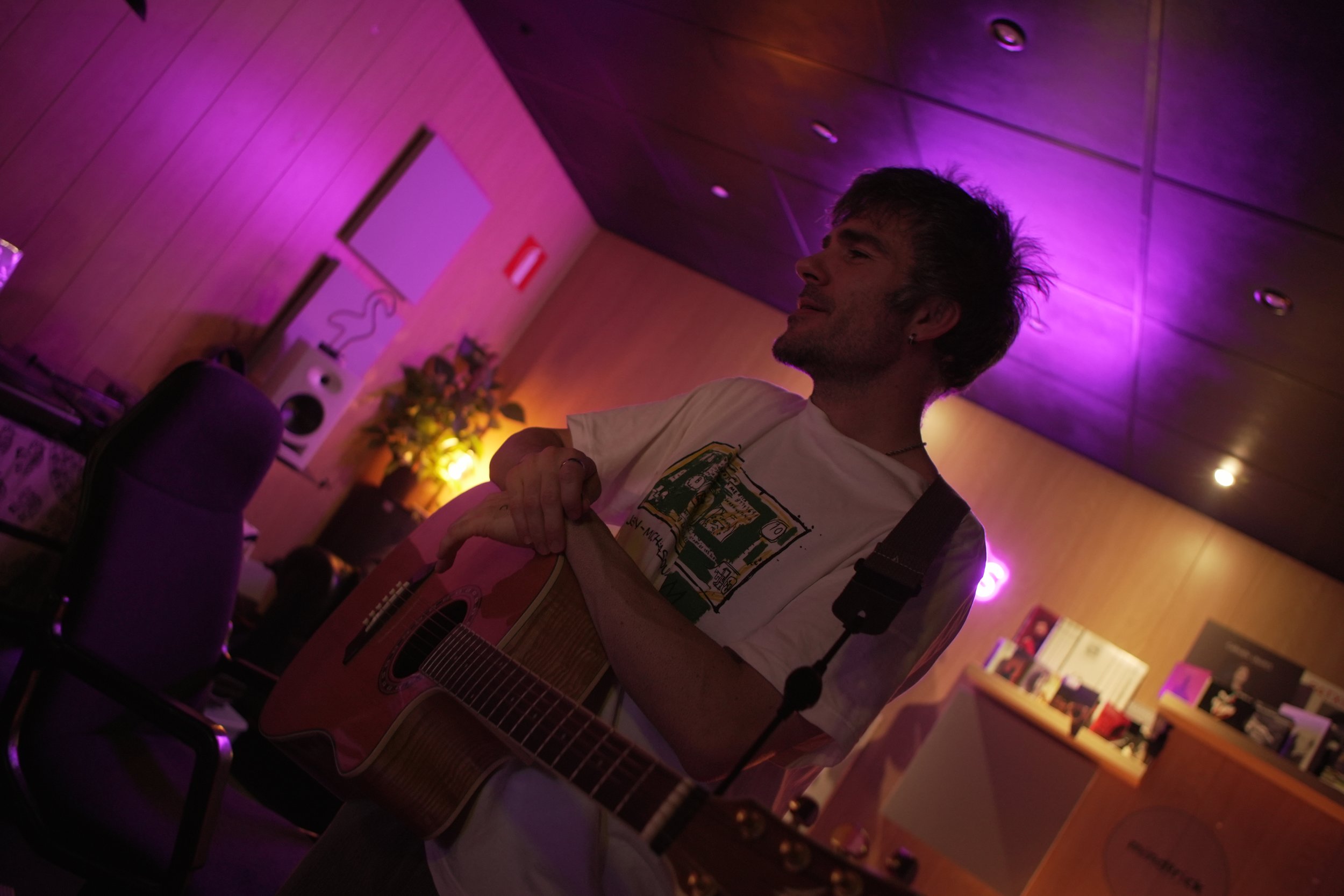 Joven tocando la guitarra en un ambiente con iluminación morada y amarilla, con paredes de madera y estanterías con libros.