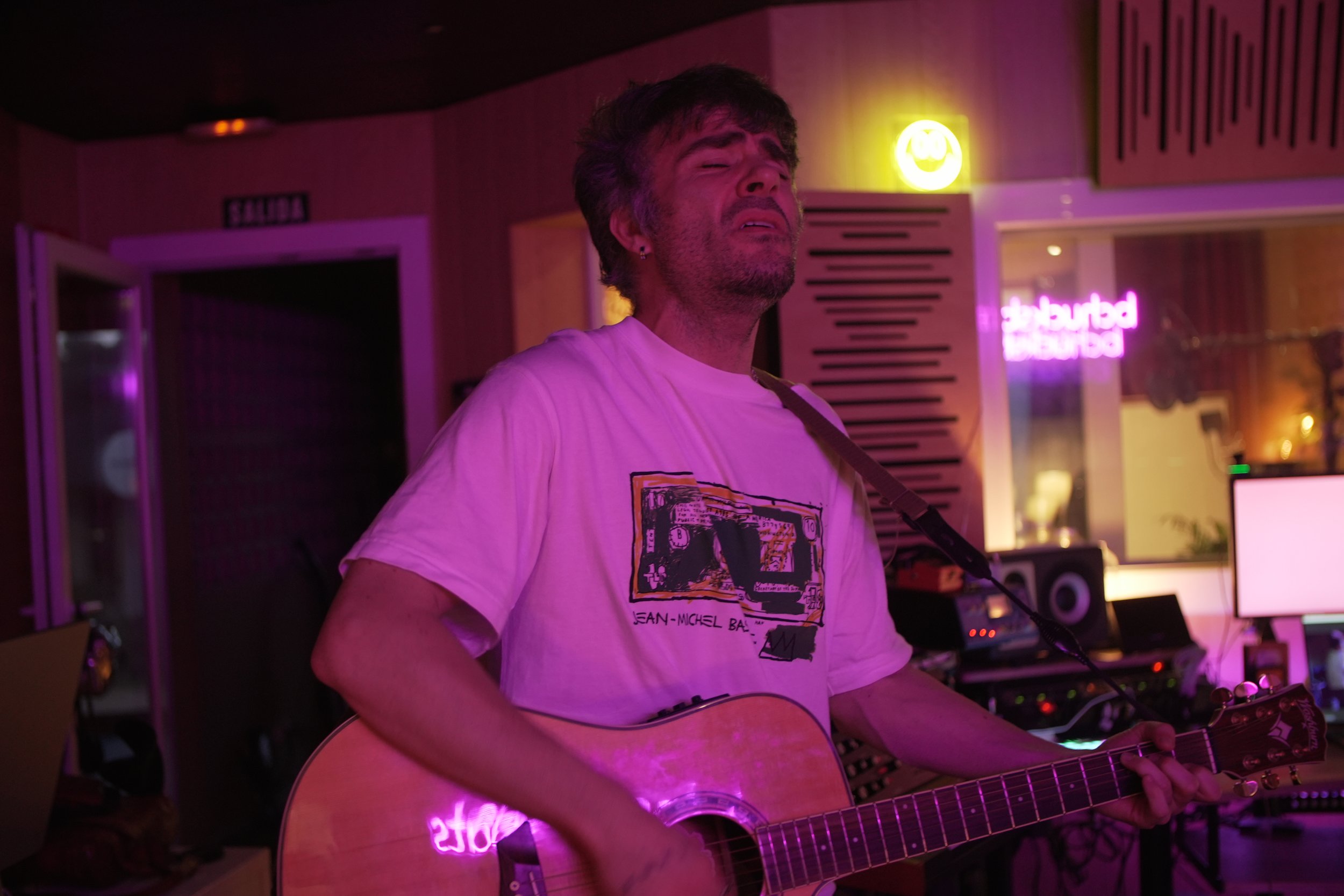 Hombre tocando guitarra en un estudio de grabación con iluminación púrpura y algunos signos y pantallas en el fondo.