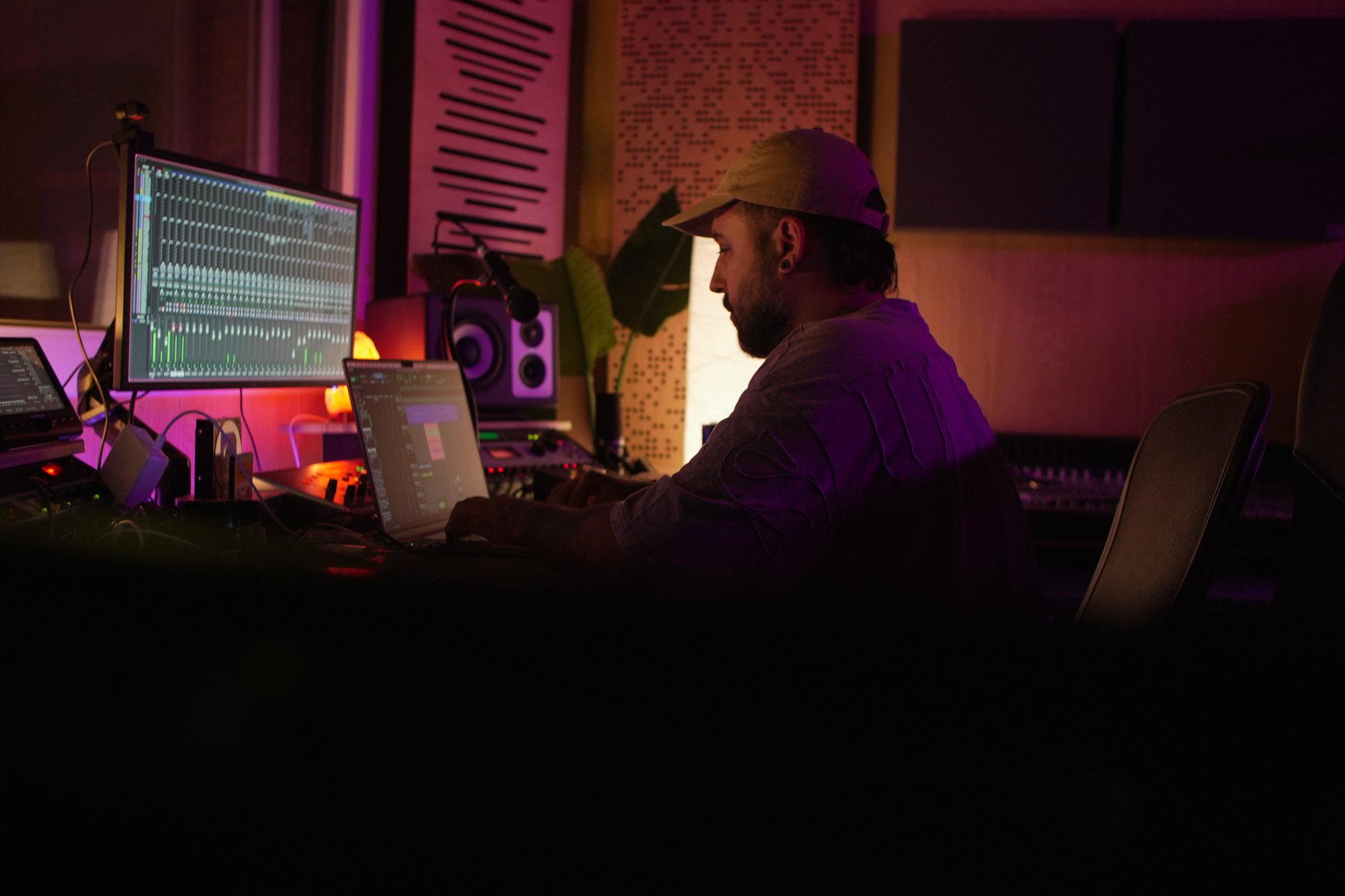 Un joven productor musical en un estudio de grabación, usando una computadora portátil y una consola de sonido, con pantallas que muestran software de mezcla y edición de audio, rodeado de equipos de estudio y cables, con iluminación tenue y ambiente