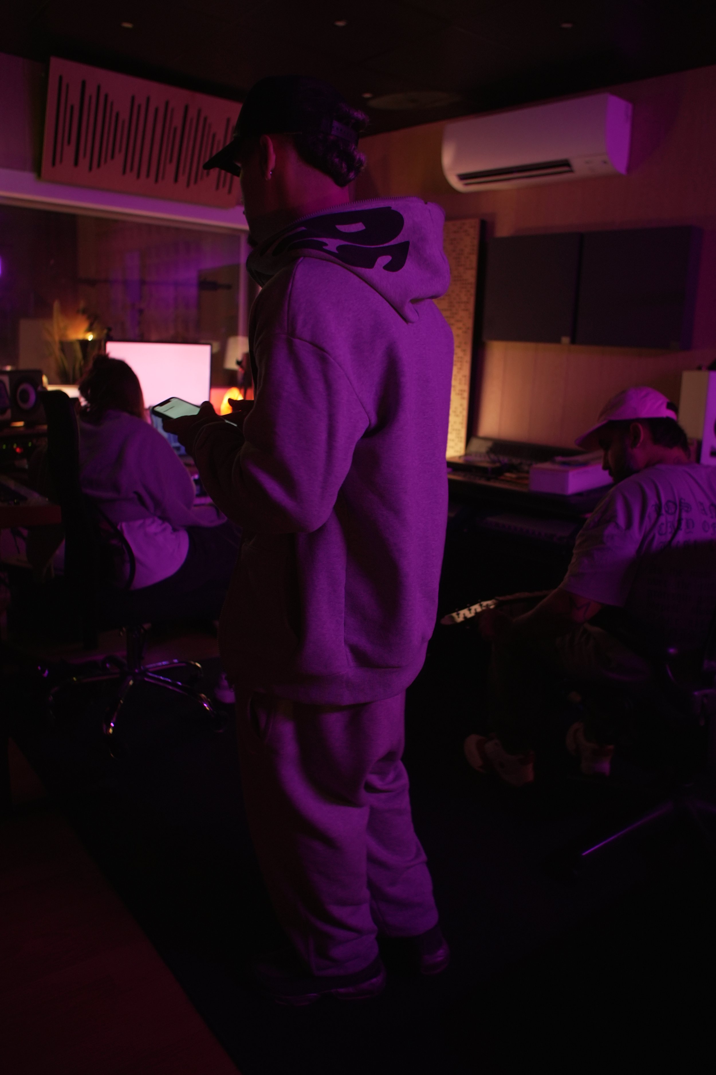 Tres personas en una sala de grabación en ambientes con luces moradas y púrpuras, una de ellas usando un teléfono móvil, otra tocando un teclado y la tercera sentada frente a una computadora.