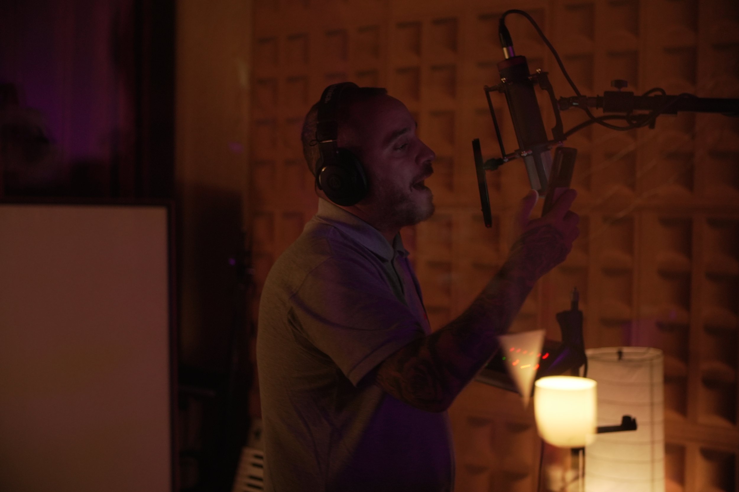 Hombre en estudio de grabación con auriculares, sosteniendo un teléfono y frente a micrófono, con ambiente oscuro y luz suave.