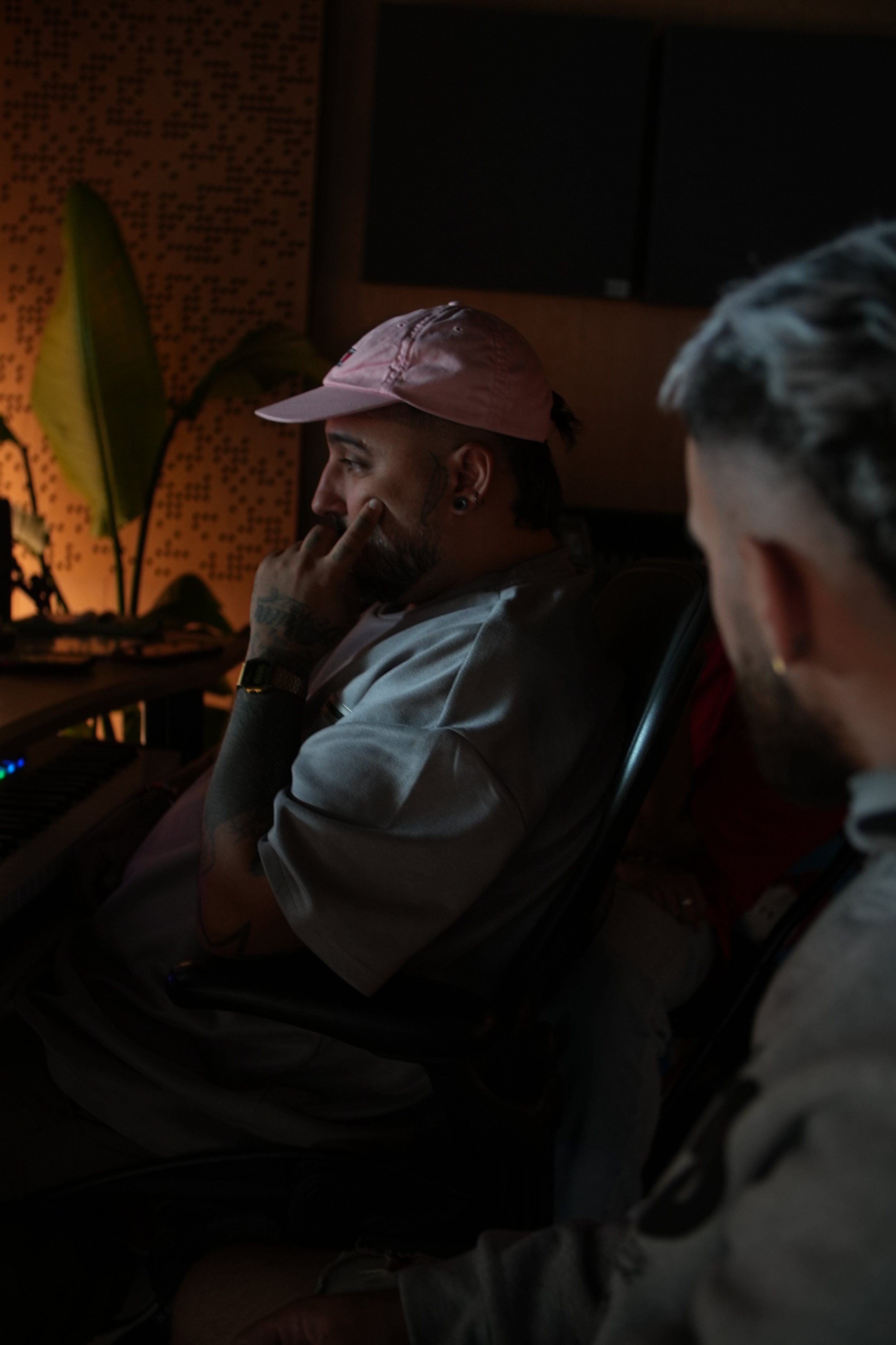 Dos hombres en un estudio de grabación, uno con gorra rosa y tatuajes, sentado frente a un equipo con pantallas oscuras detrás.