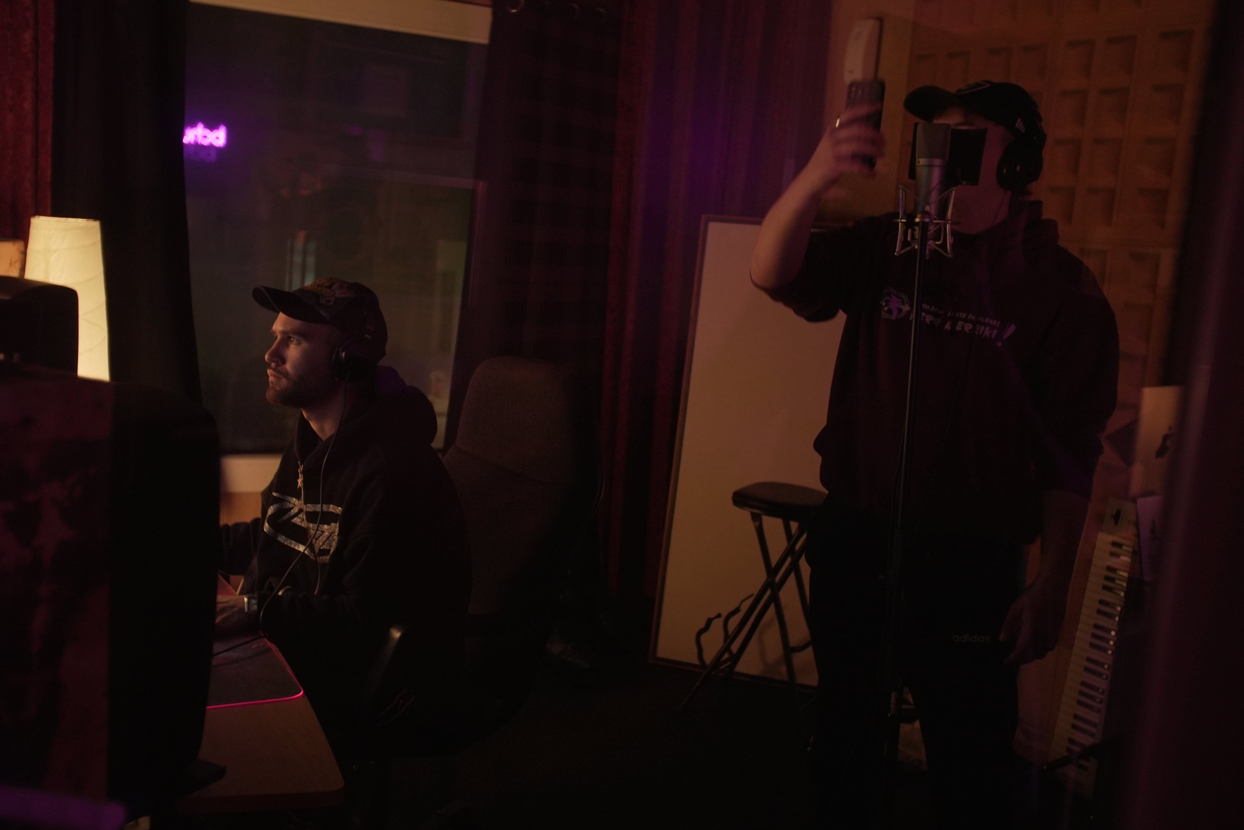 Dos personas en un estudio de grabación, una frente a una computadora con auriculares y otra con micrófono sosteniendo un teléfono, en un ambiente con iluminación tenue y equipo de grabación.