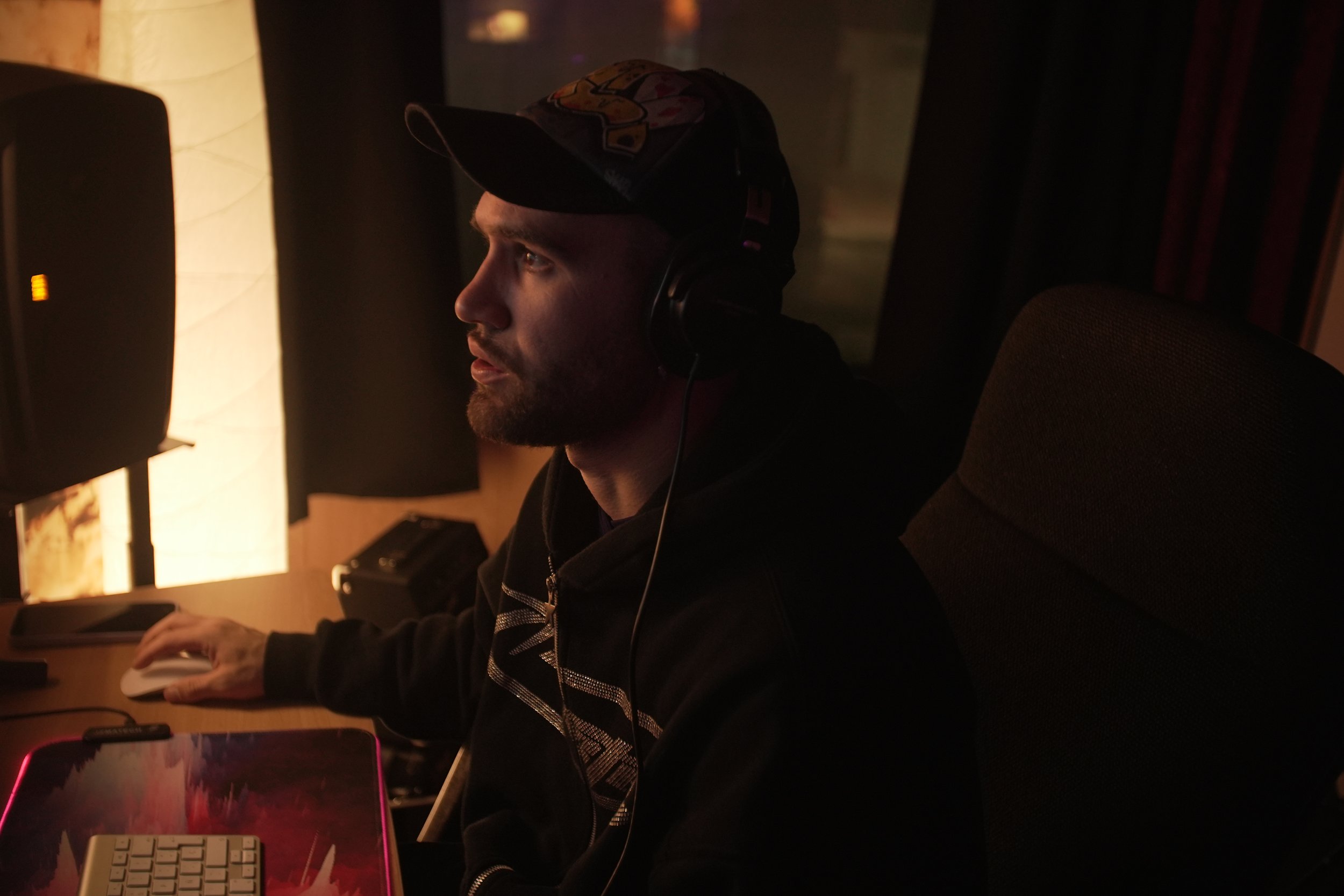 Un hombre en una cabina de grabación con auriculares, luz tenue y un computador.