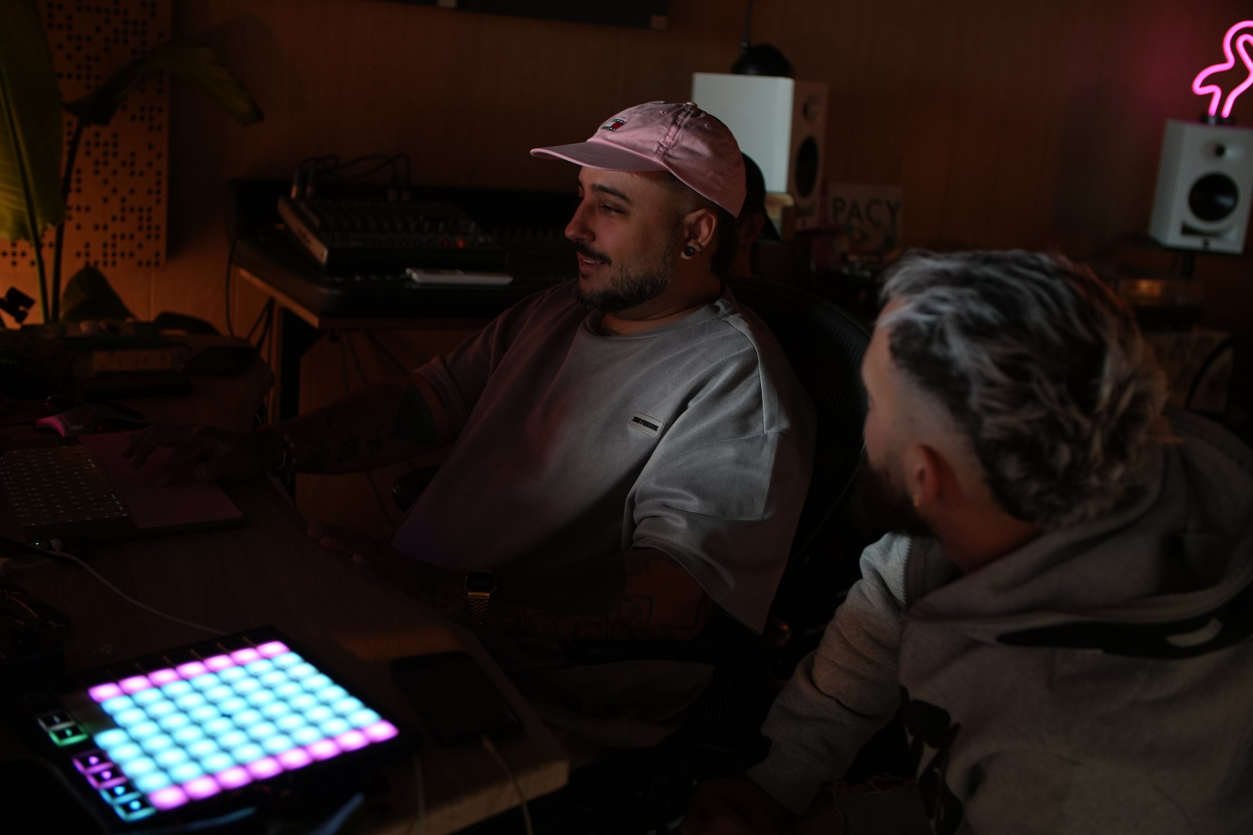 Dos personas en un estudio de grabación o producción musical, una sentado en una silla frente a un equipo de audio y la otra de pie a su lado, con sonido y luces ambientales en el fondo.