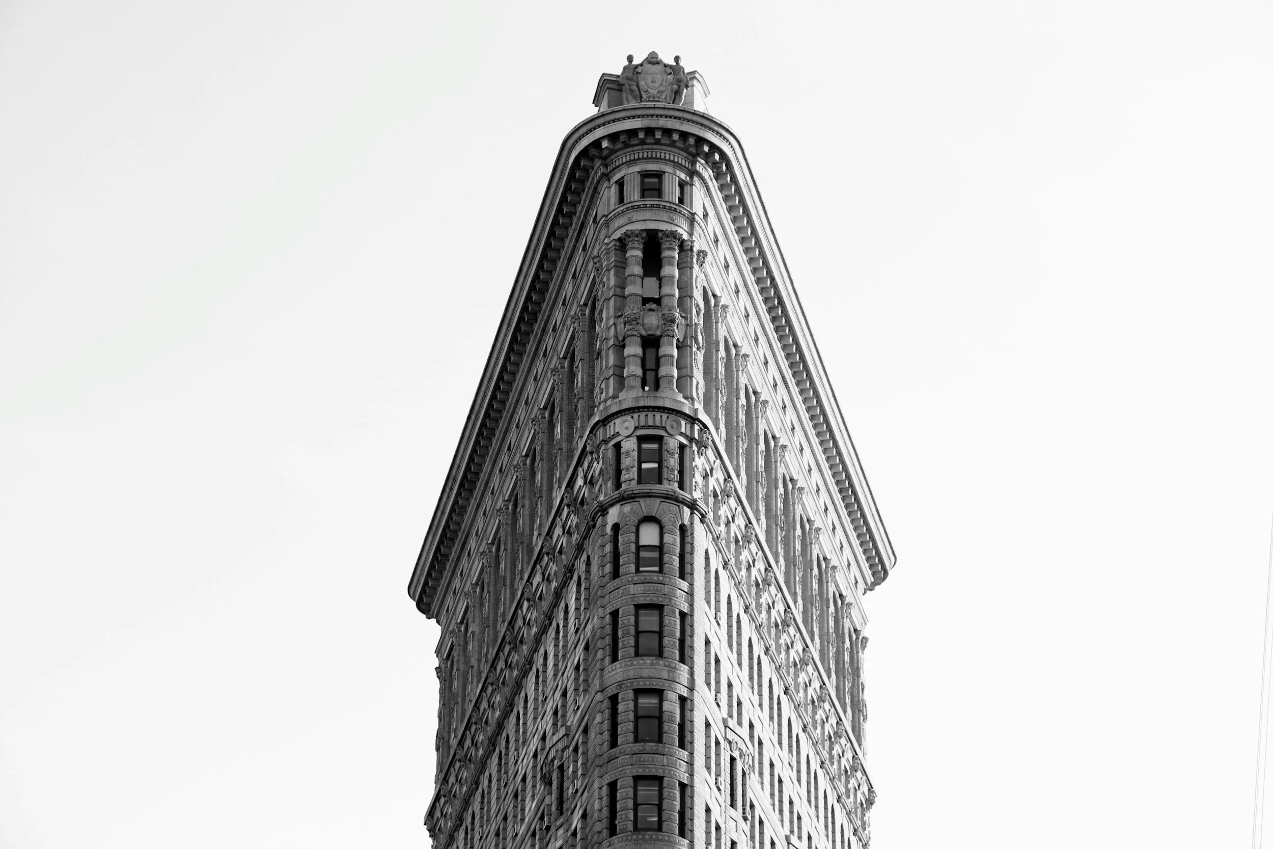 Black and white photo of the top part of a tall, historic building with intricate architectural details and sculptures on the roof.