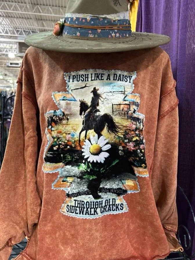 A vintage brown sweatshirt with a graphic of a person riding a bucking horse, a daisy flower, and the words 'I PUSH LIKE A DAISY THROUGH OLD SIDEWALK CRACKS'.