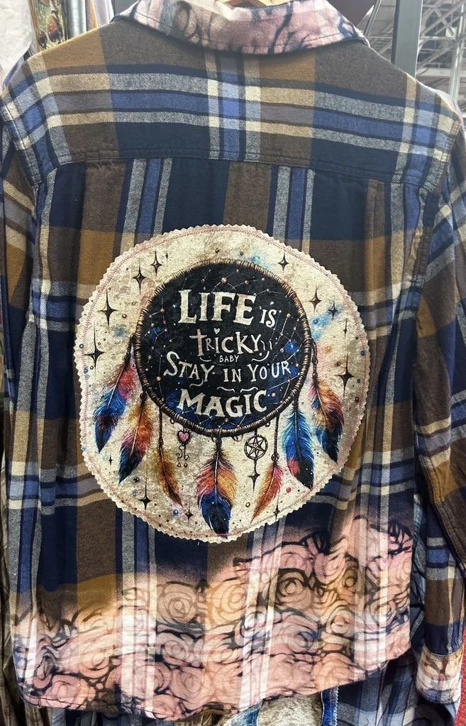 Back of a plaid jacket with a large round patch featuring a dreamcatcher and the phrase "Life is tricky baby stay in your magic."