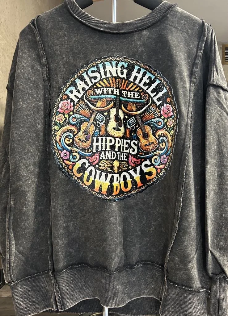 A black, vintage-style sweatshirt with colorful embroidered patch on the front. The patch shows three guitars, floral designs, and the text 'Raising Hell with the Hippies and the Cowboys' in a circular arrangement.