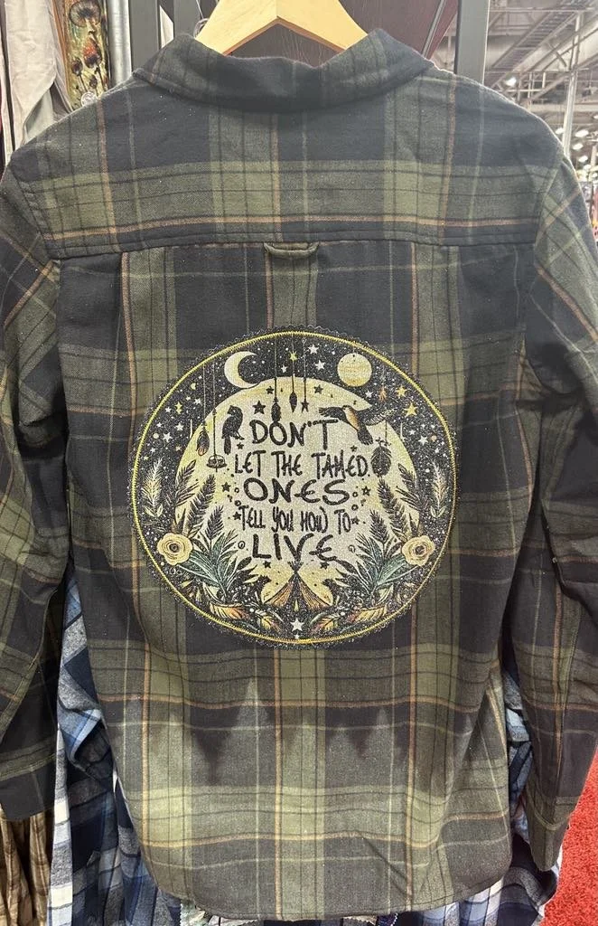 A plaid jacket with an embroidered circle design on the back featuring moons, stars, and botanicals, with the quote 'Don't let the tamed ones tell you how to live'.