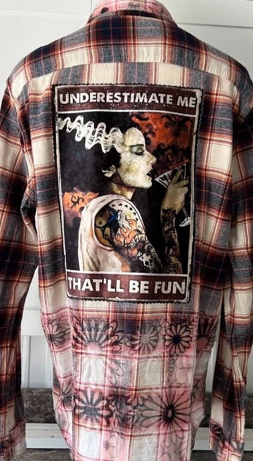 Back of a plaid shirt with a graphic patch of a woman's profile holding a cigarette, with the text "Underestimate Me, That'll Be Fun."