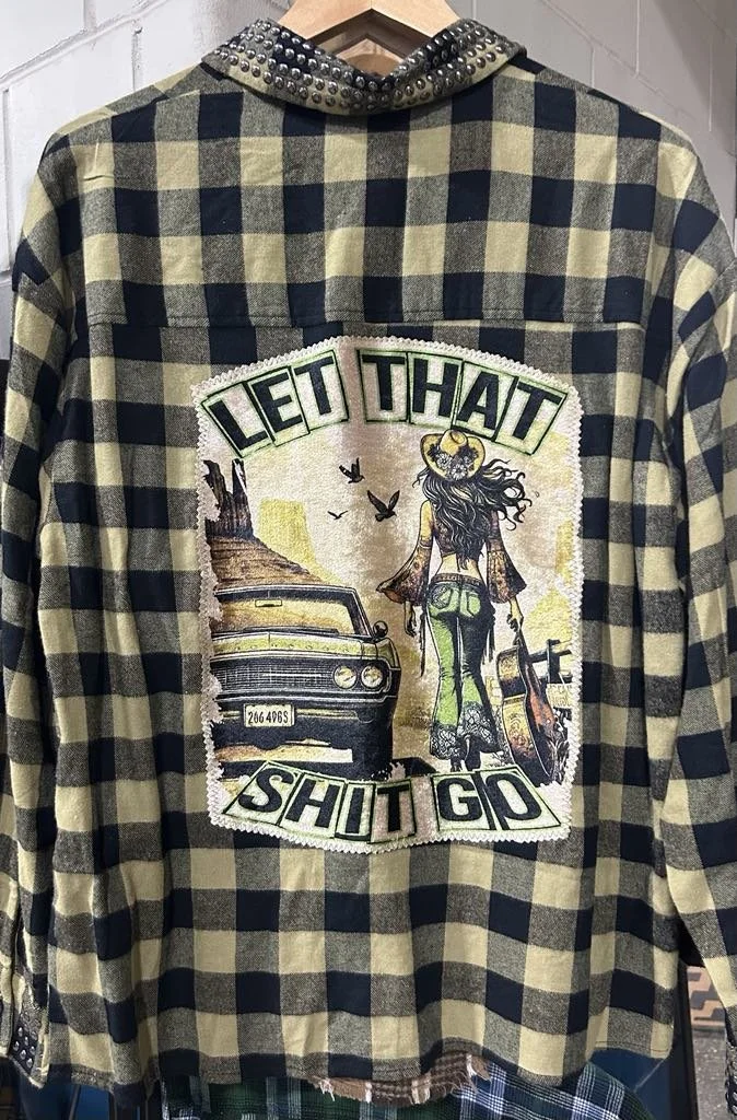 A plaid flannel shirt with a large embroidered patch on the back. The patch depicts a girl with long hair wearing a wide-brimmed hat, walking away from an old car in a desert landscape with rock formations and birds. The patch has the phrase "Let That Shit Go" embroidered on it.