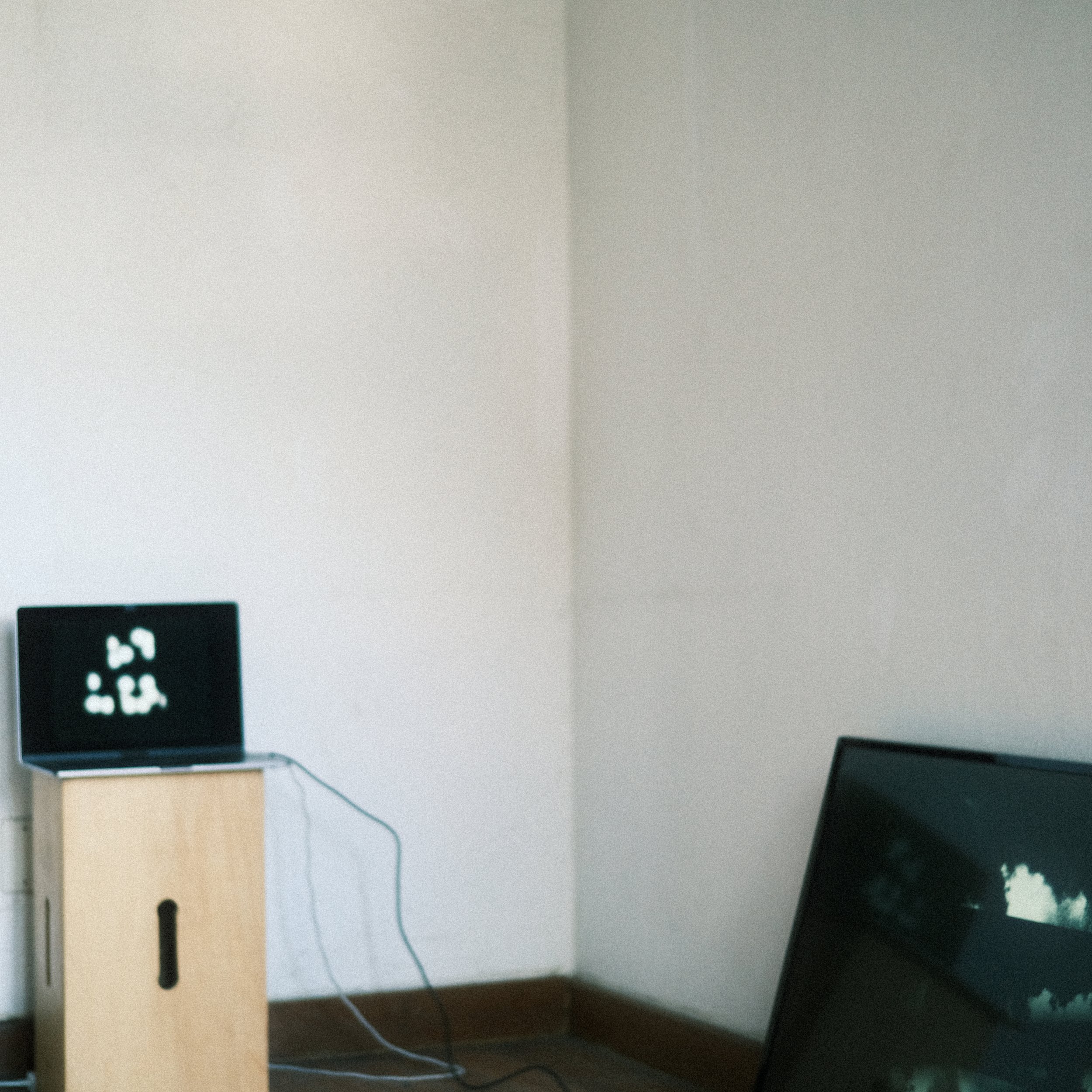 Estação de trabalho com duas telas, uma pequena em uma mesa de madeira ao lado da parede, exibindo elementos gráficos em branco sobre fundo preto, e uma maior tela de TV ou monitor parcialmente visível ao lado, também exibindo elementos gráficos.