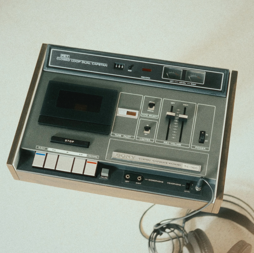 Fita cassete antiga conectada a um gravador de fita de áudio com controles, botões e cabos, além de um par de fones de ouvido ao lado.
