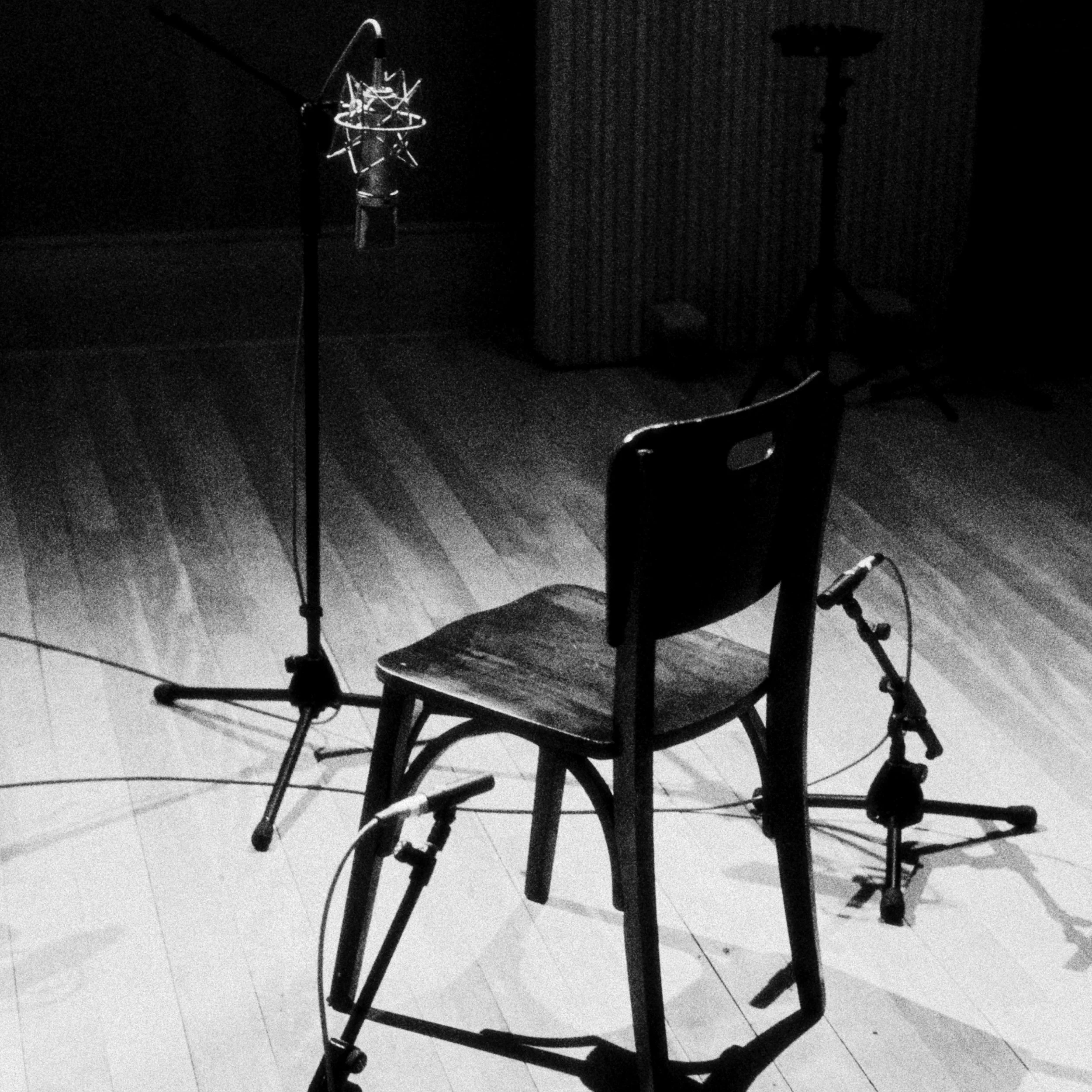 Cadeira de madeira no centro de um palco, com microfones e suportes ao redor, iluminação focada nela, fundo escuro.