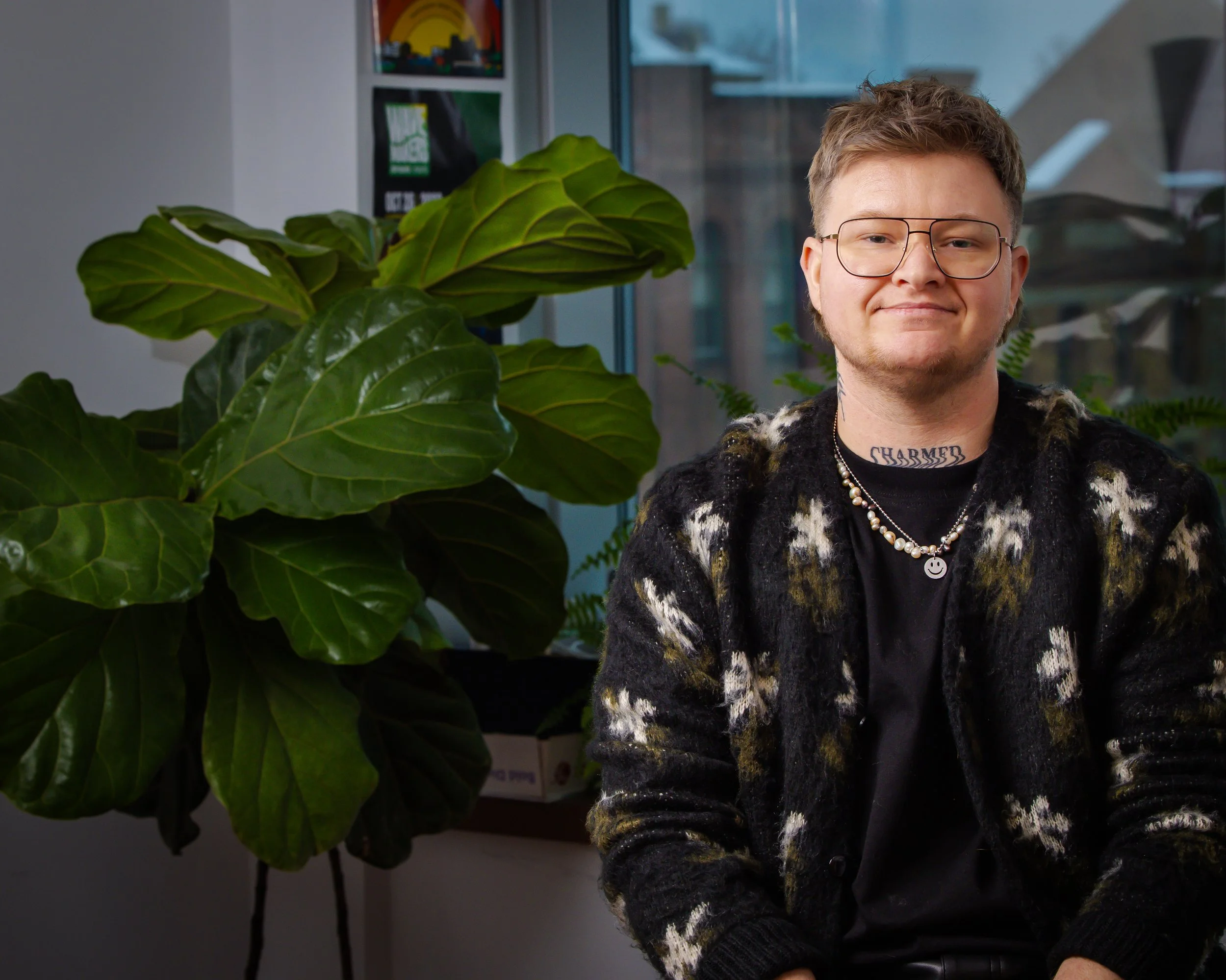 Sits near a sunlit window, accompanied by a thriving fiddle-leaf fig plant. They wear a black cardigan adorned with woolly white flowers and green leaves, paired with classic aviator glasses. Their strong, friendly smile is warm and inviting.