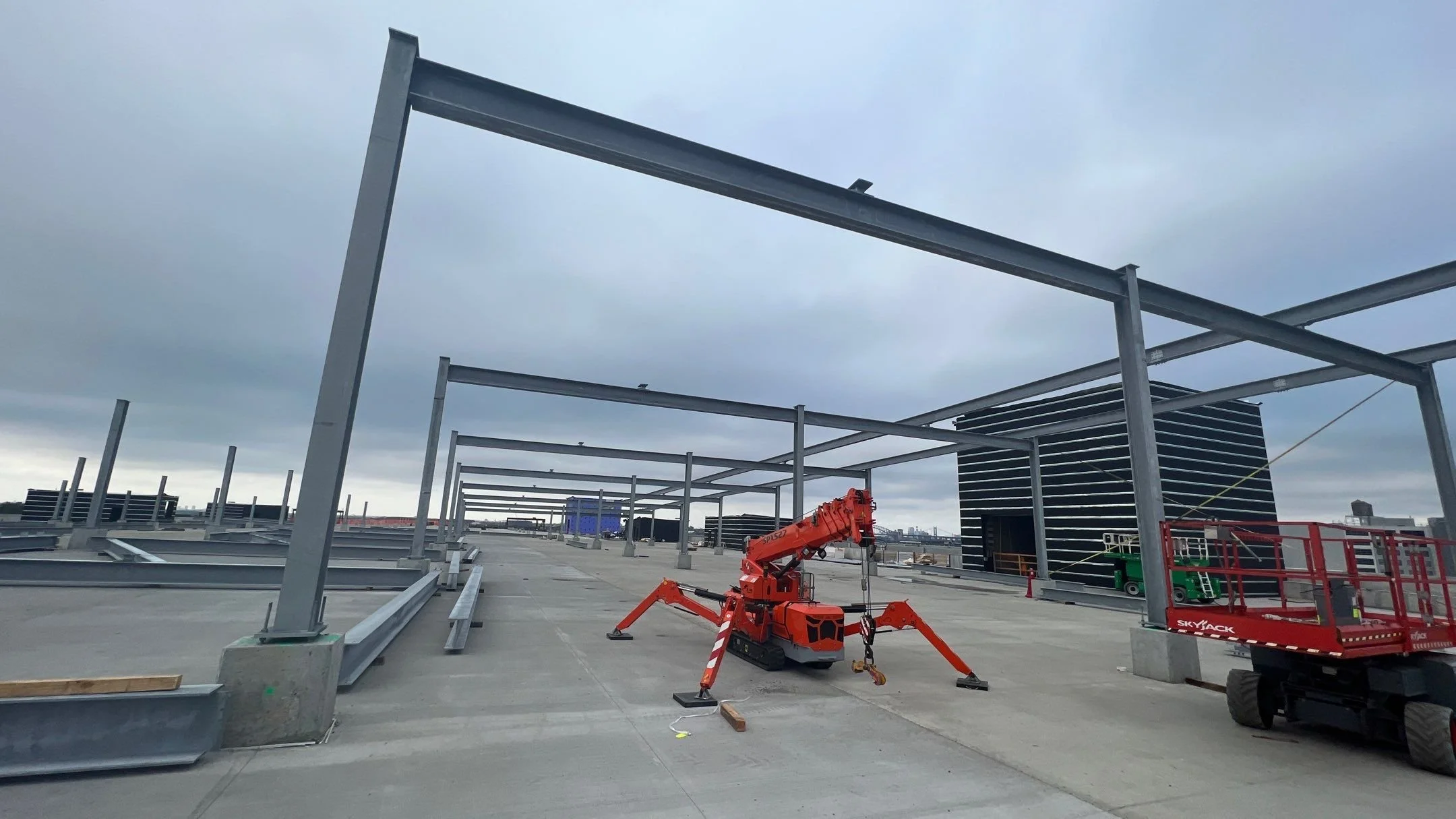 Construction site on a building rooftop with metal framework, a small red crane, and construction equipment under cloudy skies.