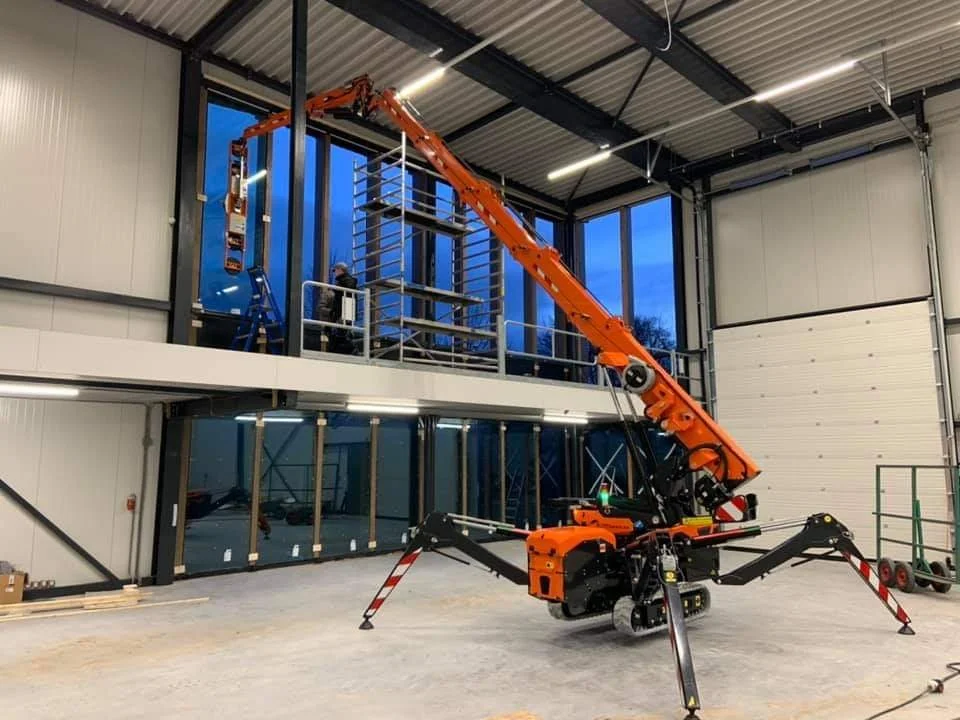 An orange and black robotic crane working inside a modern building, lifting a metal shelf to a second-floor balcony or mezzanine.