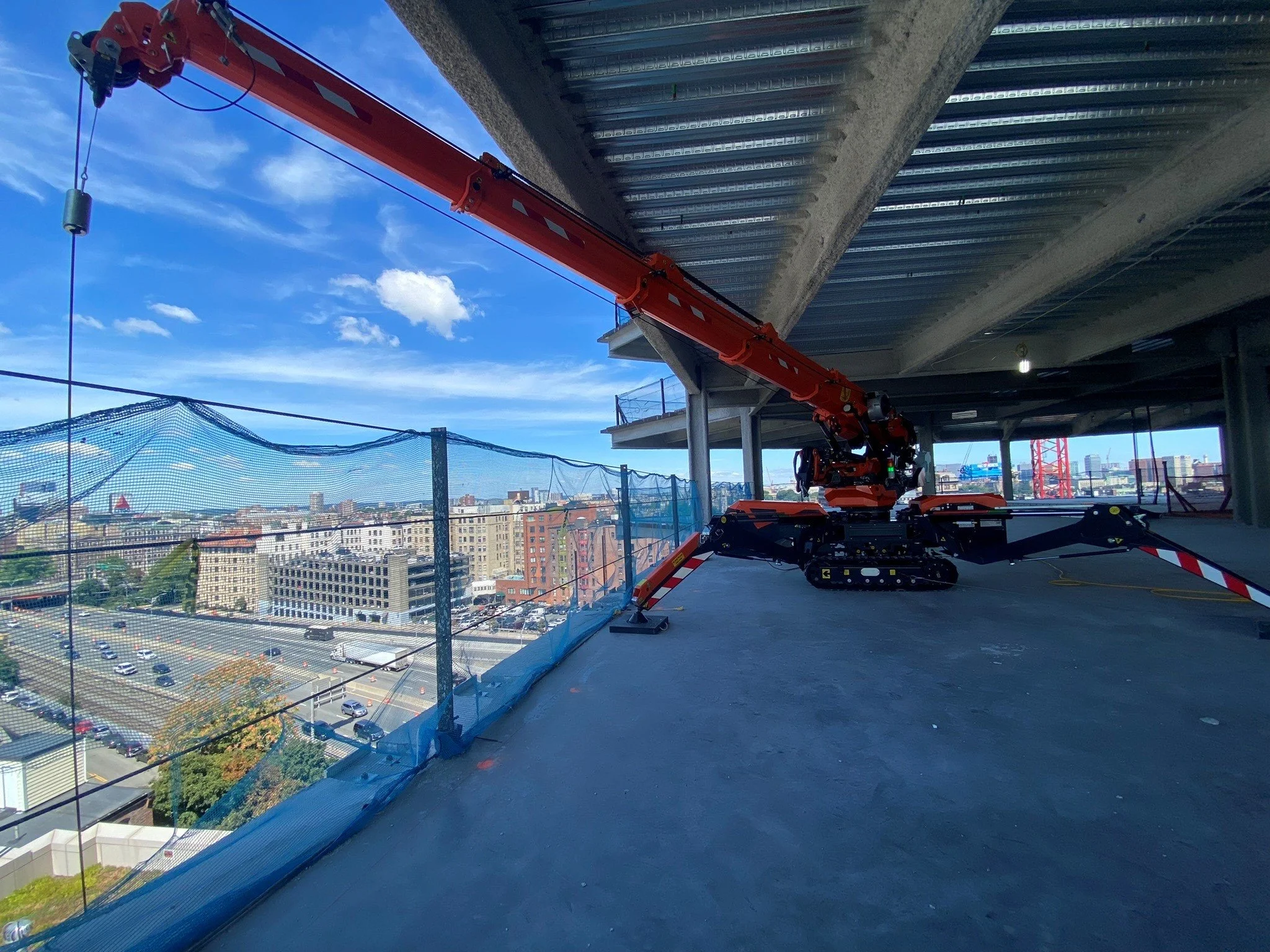 Construction site on a high-rise building with a large orange crane and cityscape view, blue sky, and some clouds.