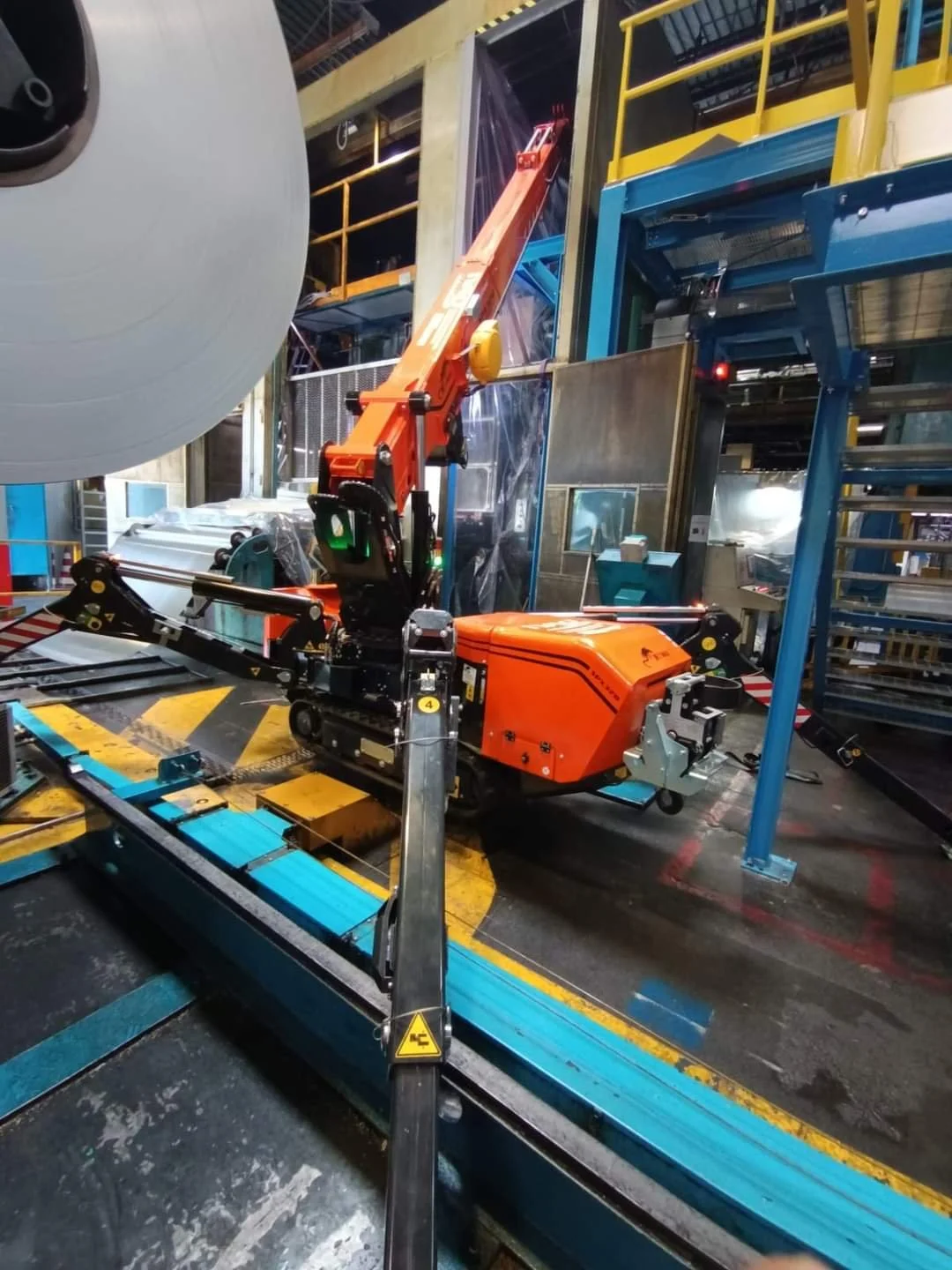 An orange robotic arm working in an industrial manufacturing setting, surrounding large steel rolls and machinery.