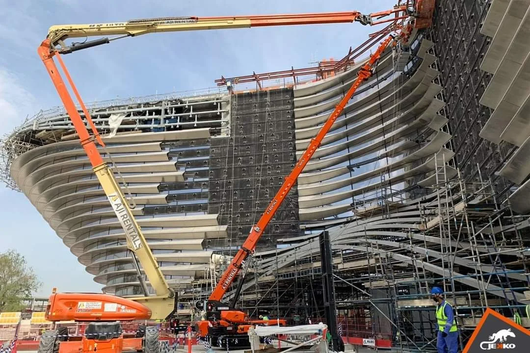 Construction workers and cranes working on a modern building with curved, layered architecture and scaffolding
