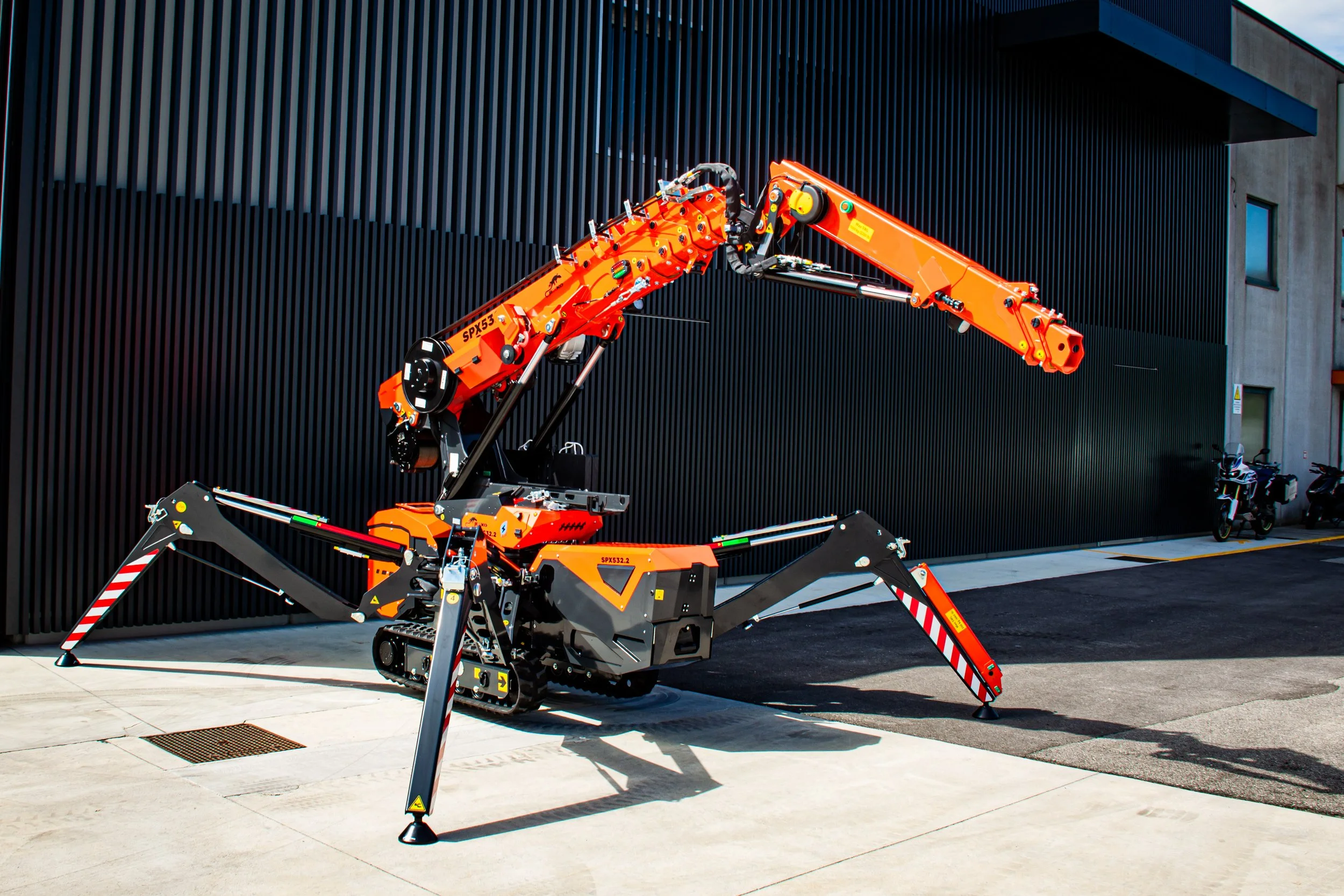 A large, orange robotic arm with black and yellow accents, situated outdoors near a modern building with black vertical siding.
