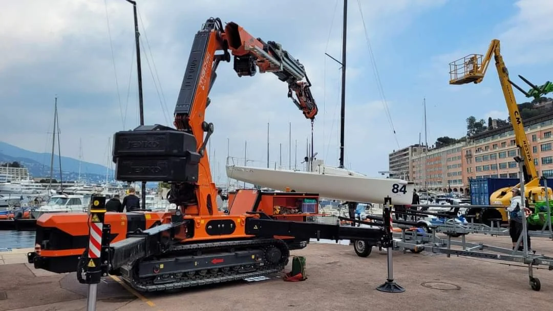 A large orange crane lifting a white sailboat with the number 84 on it near a marina, with other boats and buildings in the background.