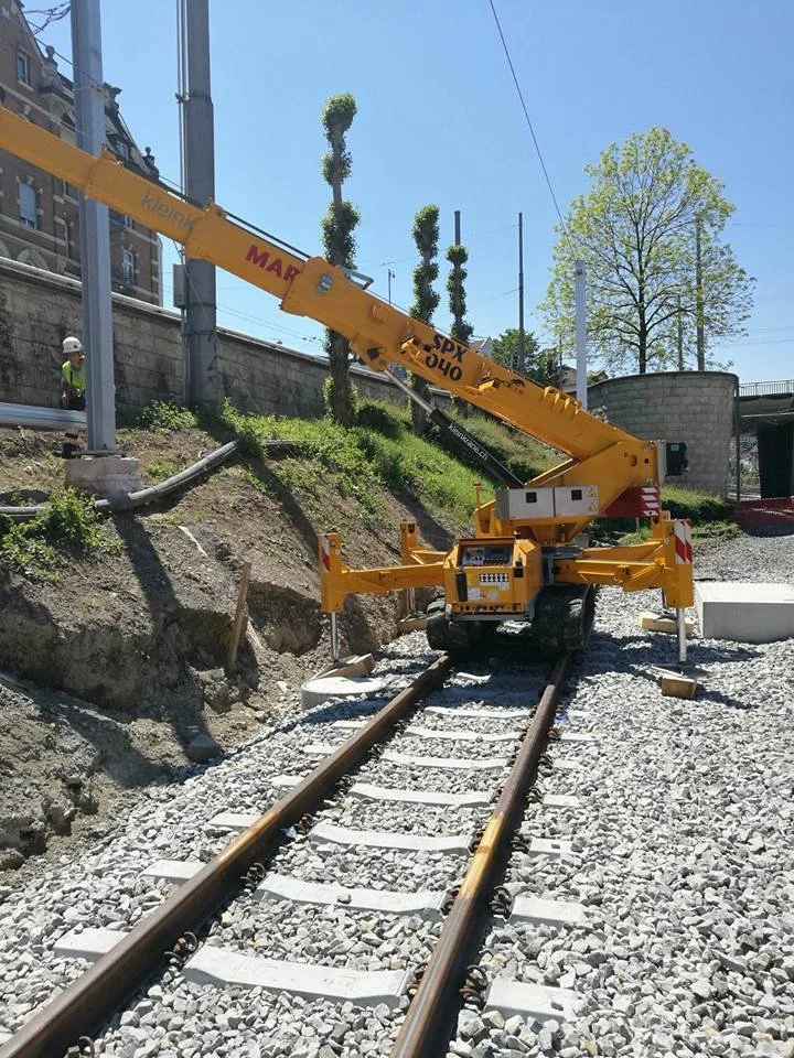 A yellow rail construction vehicle is working on a railway track, with its arm extended over the tracks and a worker nearby observing the site on a sunny day.
