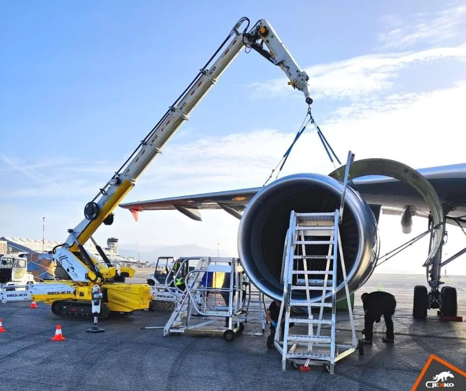 Workers using a crane to lift a jet engine onto an airplane at an airport tarmac during daytime.