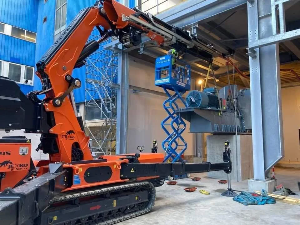 Construction site with an orange crane lifting materials. The crane is positioned near a partially constructed building with steel beams and scaffolding. Workers' safety equipment and tools are visible on the ground.