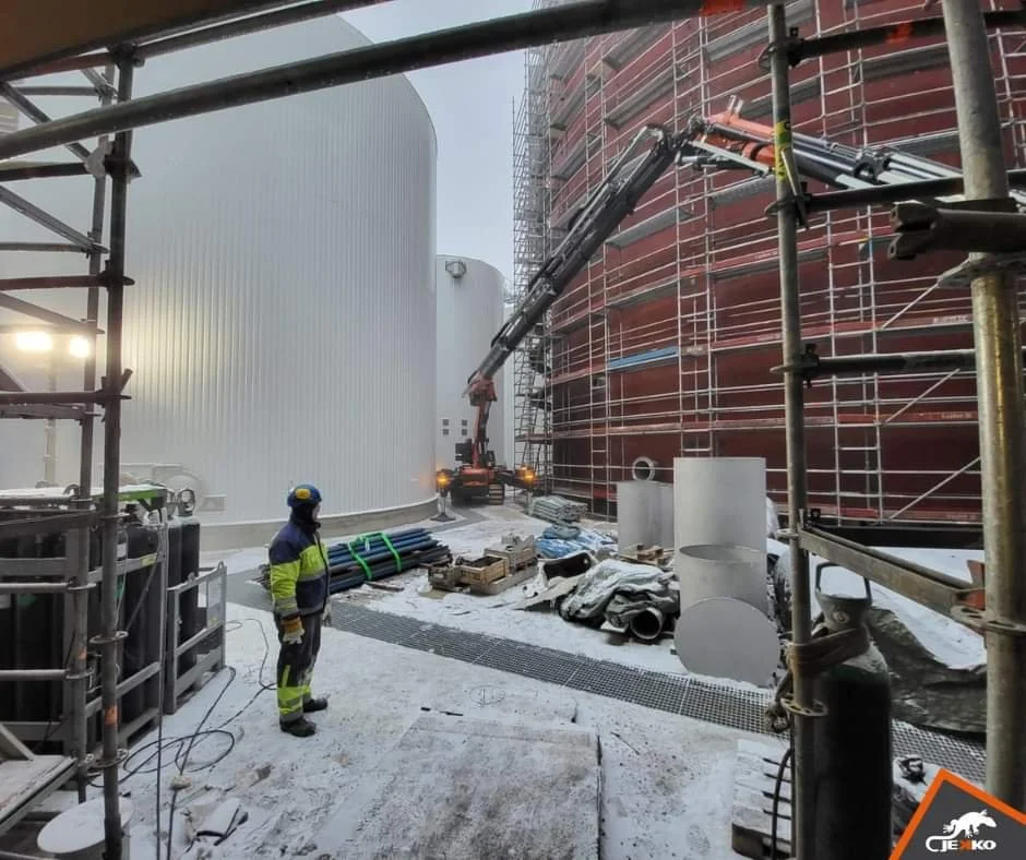Construction site with scaffolding, a worker in safety gear, and a crane lifting materials on a snowy day.
