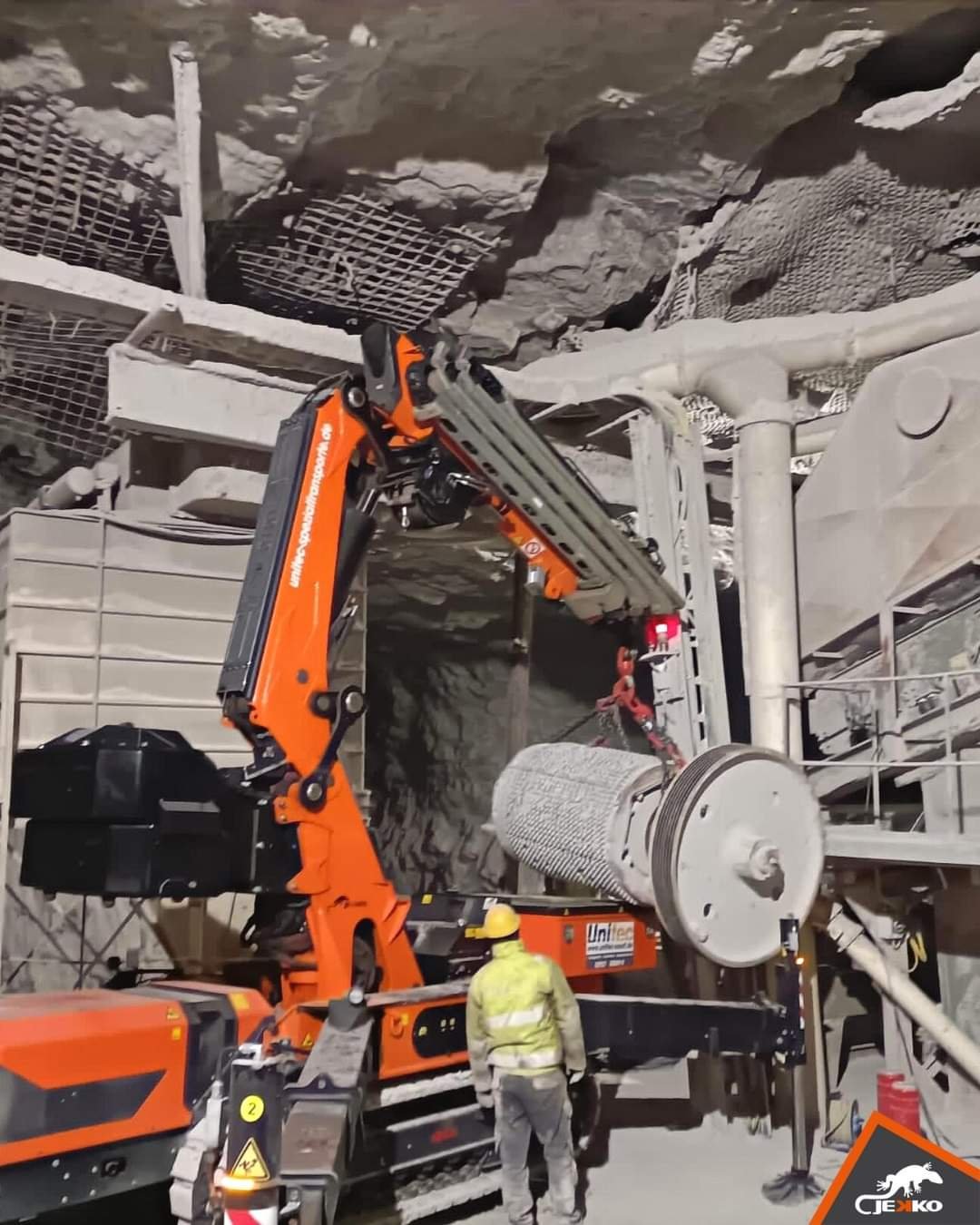 A construction worker in a yellow hard hat and reflective vest standing next to an orange hydraulic drilling machine inside an underground mine or tunnel with rocky ceiling and metal safety structures.