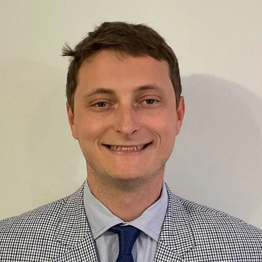 A young man with short brown hair, smiling, wearing a checkered blazer, a light blue dress shirt, and a dark blue tie, standing against a white wall.